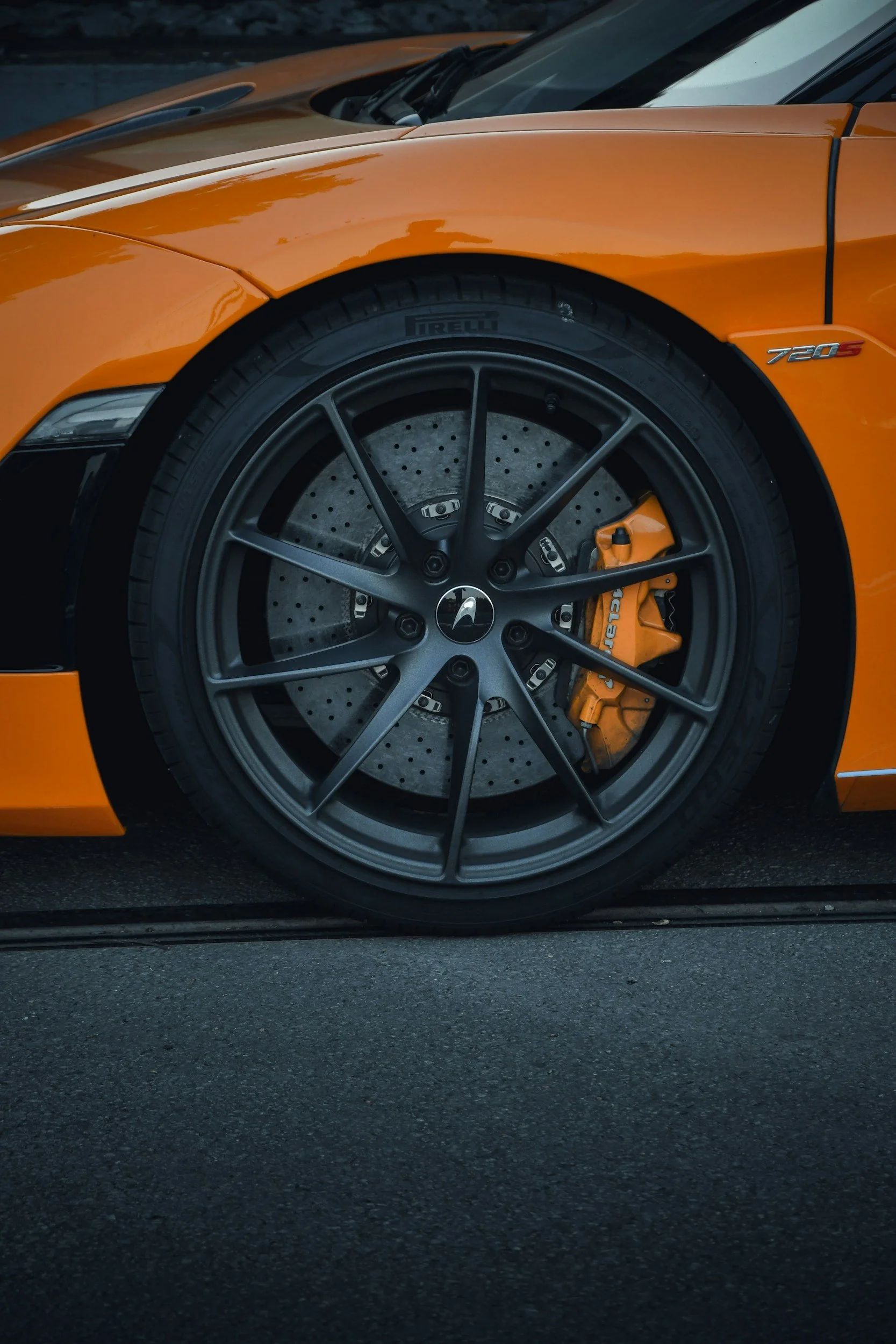 Ein orangefarbener Sportwagen mit schwarzem Felgendesign, einem großen Bremssattel und perforiertem Bremsscheibe, auf einer dunklen Asphaltstraße.
