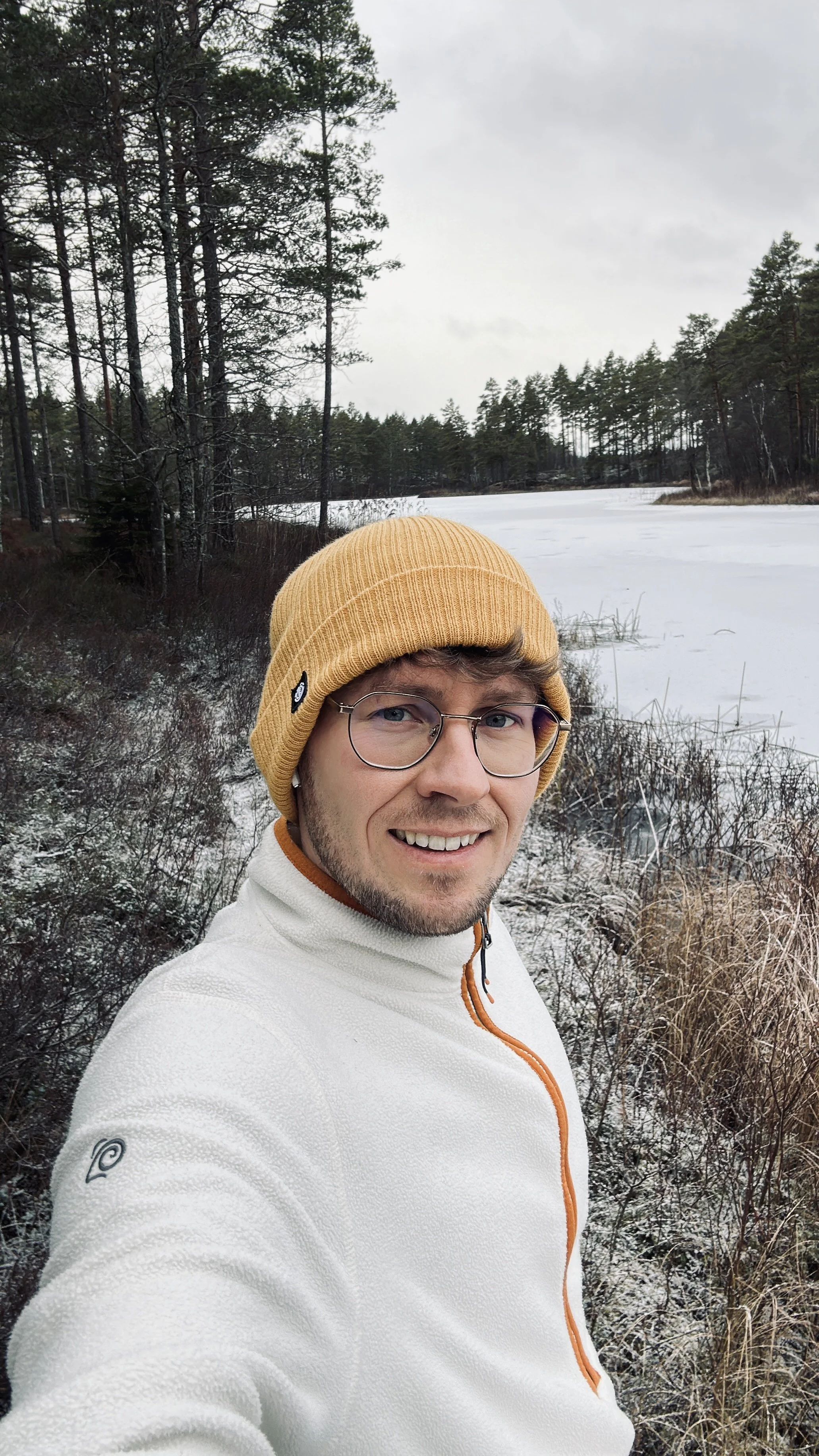 Ein lächelnder junger Mann trägt eine gelbe Mütze und eine weiße Jacke mit einem orangefarbenen Reißverschluss, steht vor einer verschneiten Winterlandschaft mit Bäumen und einem zugefrorenen See.