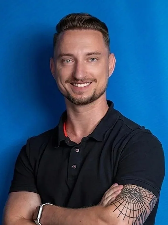 Ein junger Mann mit kurzen, glatt frisierten Haaren, einem gepflegten Bart und einem Tattoo am rechten Arm, trägt ein schwarzes Polo-Shirt vor einem blauen Hintergrund.