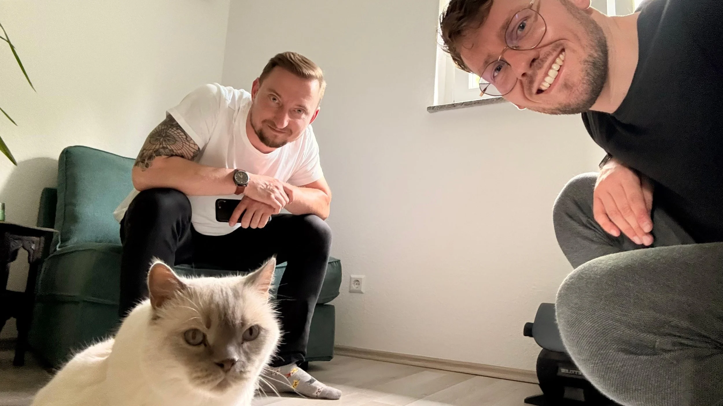 Zwei lächelnde Männer sitzen im Wohnzimmer, vor ihnen eine weiße Katze mit hellen Augen. Der Mann auf der linken trägt ein weißes T-Shirt und hat Tattoos am Arm, der Mann auf der rechten trägt eine schwarze Kleidung und trägt Brille.