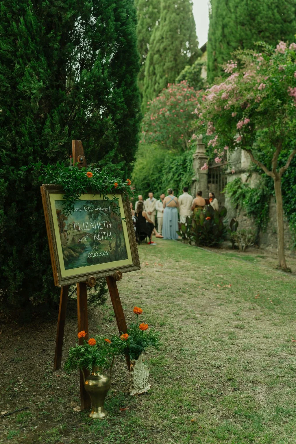 Wedding welcome sign on a vintage painting with greenery and orange flowers leading to guests gathered in a garden setting.