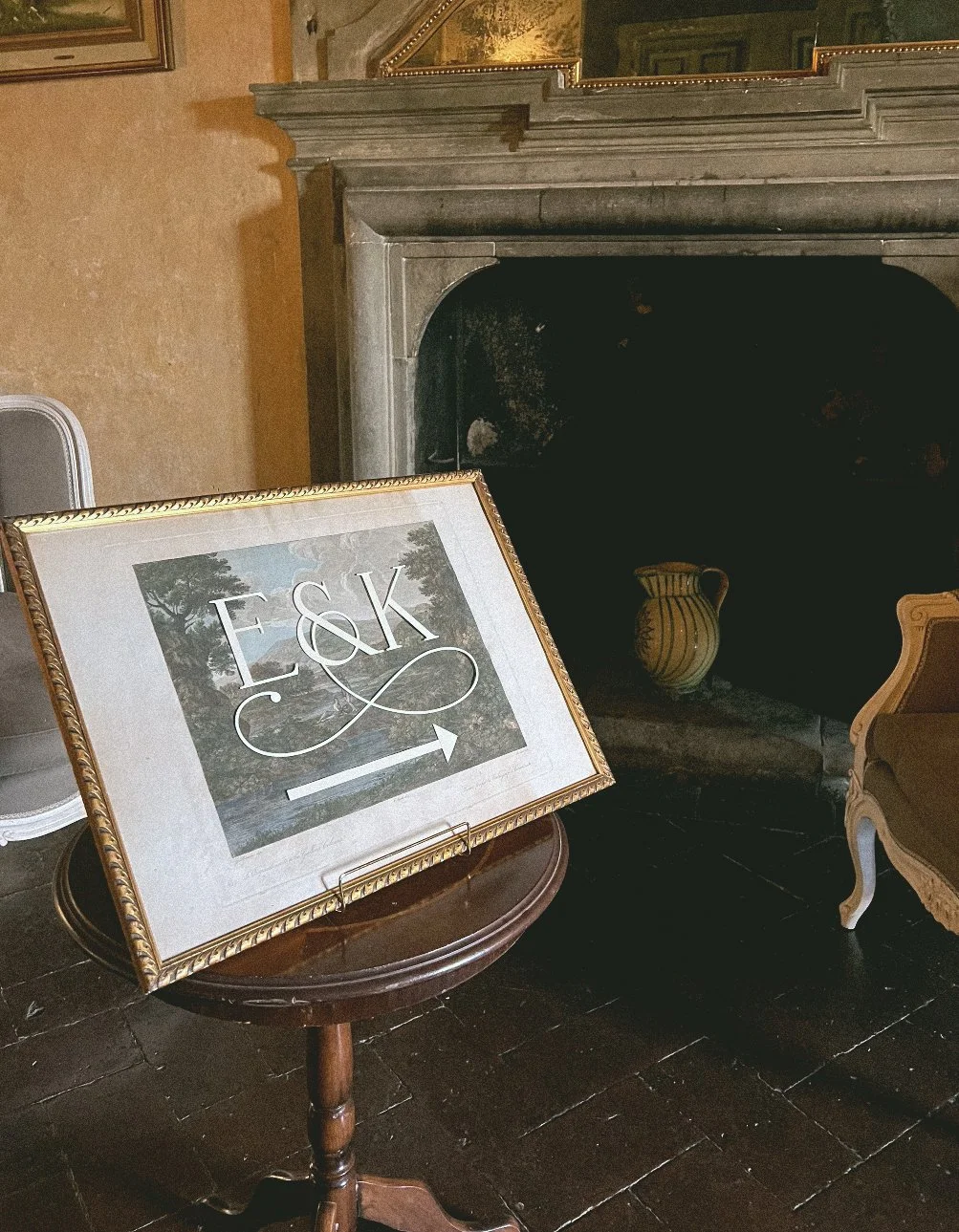 A framed wedding welcome sign on a vintage artwork with the initials E & K and an arrow pointing to the right, placed on a small wooden table in front of a dark fireplace at a castle with beige pottery inside.