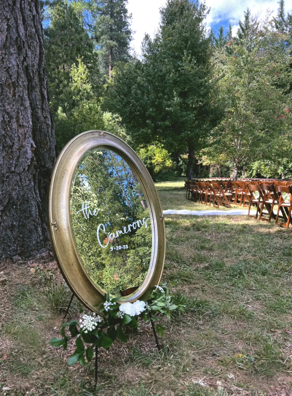 An oval mirror welcome sign with ornate gold frame and white flowers at the base, reflecting trees and sky, set outdoors next to a large tree, with rows of wooden chairs arranged on the grass, indicating a wedding venue or outdoor ceremony setup.