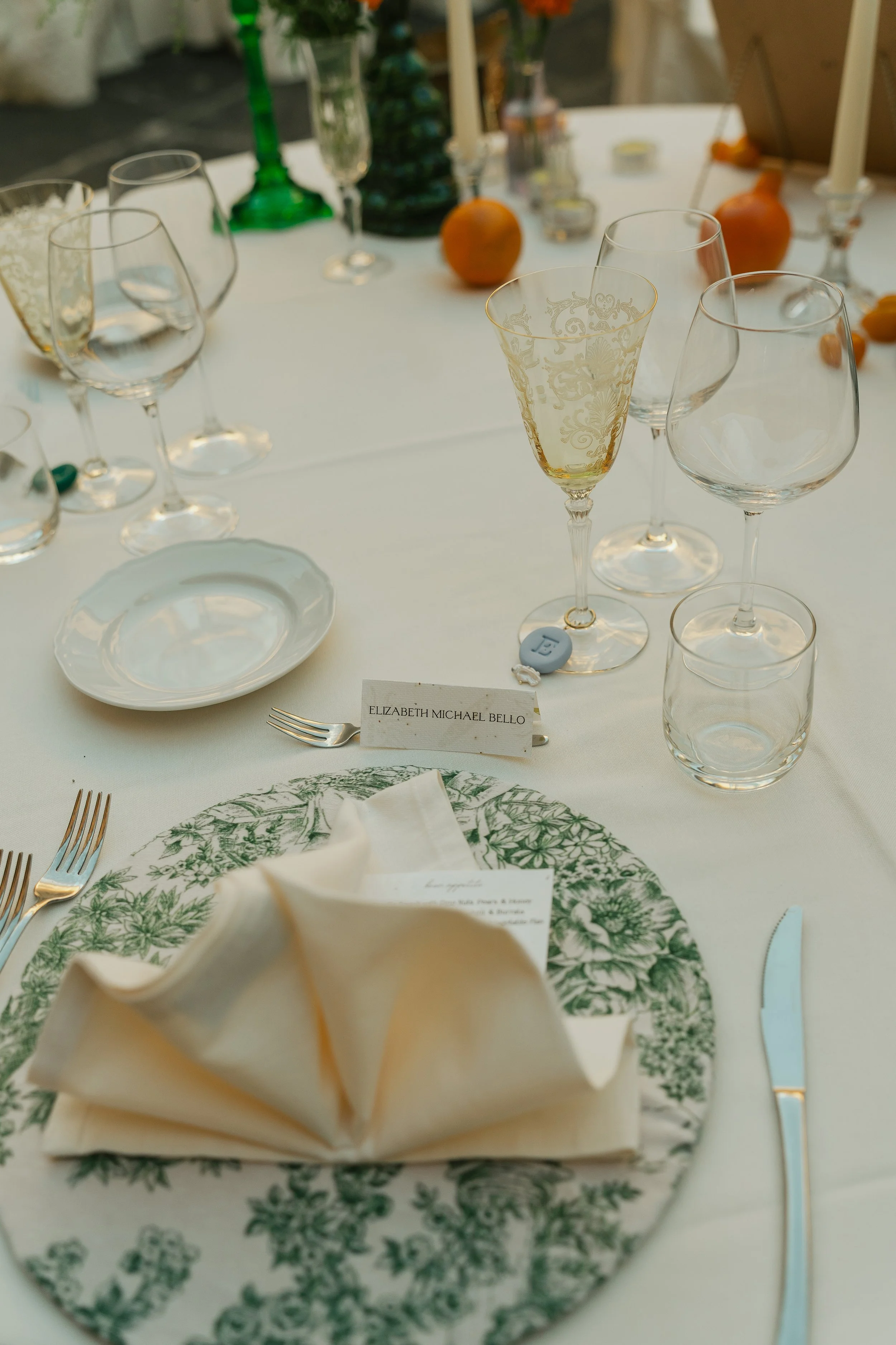 A table set for a formal wedding event with wine glasses, a plate with a green chinoiserie pattern, a folded napkin, & cutlery. There are name cards on seed paper. The background features additional vintage glassware, candles, & decorative elements.