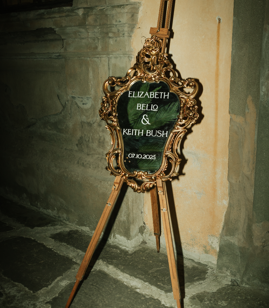 A decorative vintage wedding sign on a wooden easel with gold ornate frame, displaying bride and groom names on an antique styled mirror, against a stone and plaster wall.