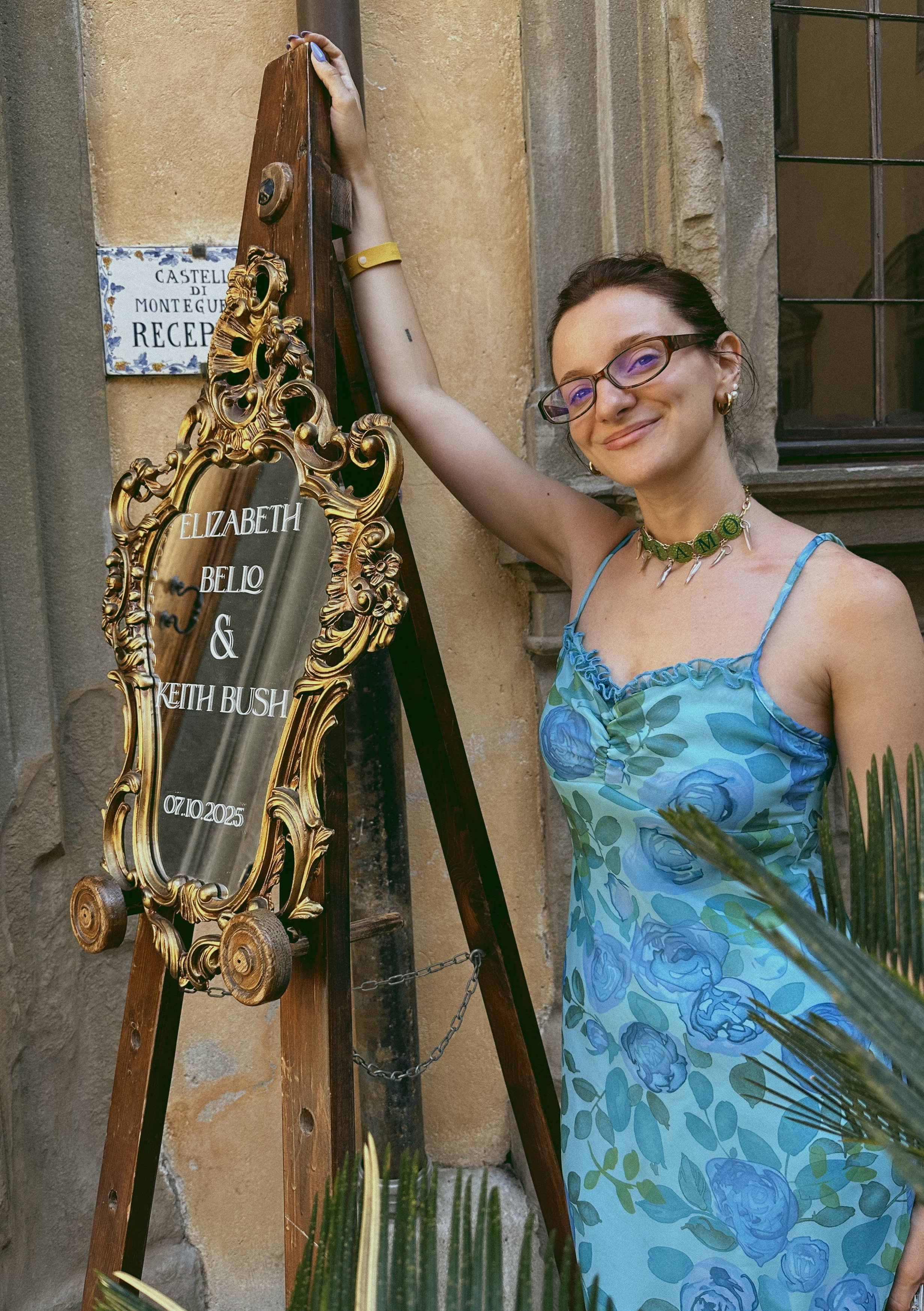 A smiling woman with glasses, wearing a blue dress with a floral pattern, standing next to a decorative gold-framed mirror and a wooden easel with a sign that reads "Elizabeth Bello & Keith Bush, 07.10.2025." She is standing outside near a stone building, with some plants in the foreground.