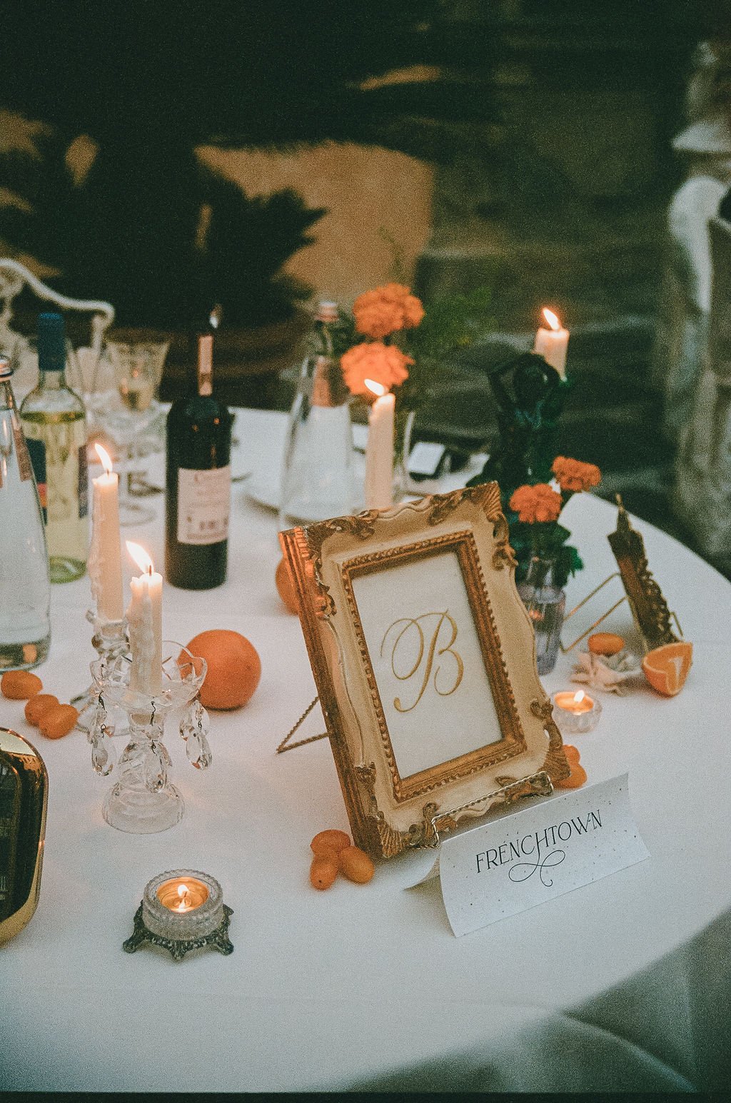 A decorated table with lit candles, orange flowers, a framed monogrammed sign with the letter B, bottles of wine, and orange slices, set for a celebration or dinner party.