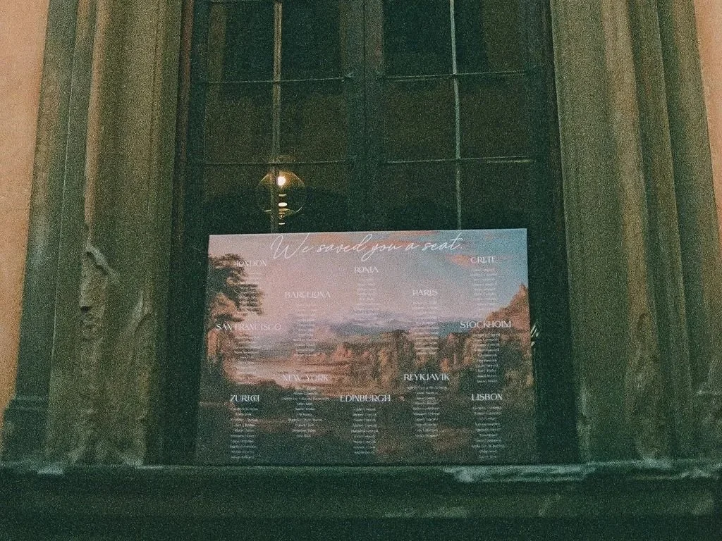 A large seating chart photo display board titled "We saved you a seat" showing a vintage painting with country names and cities listed as table names, mounted on a stone wall inside a dimly lit courtyard wedding in Italy.