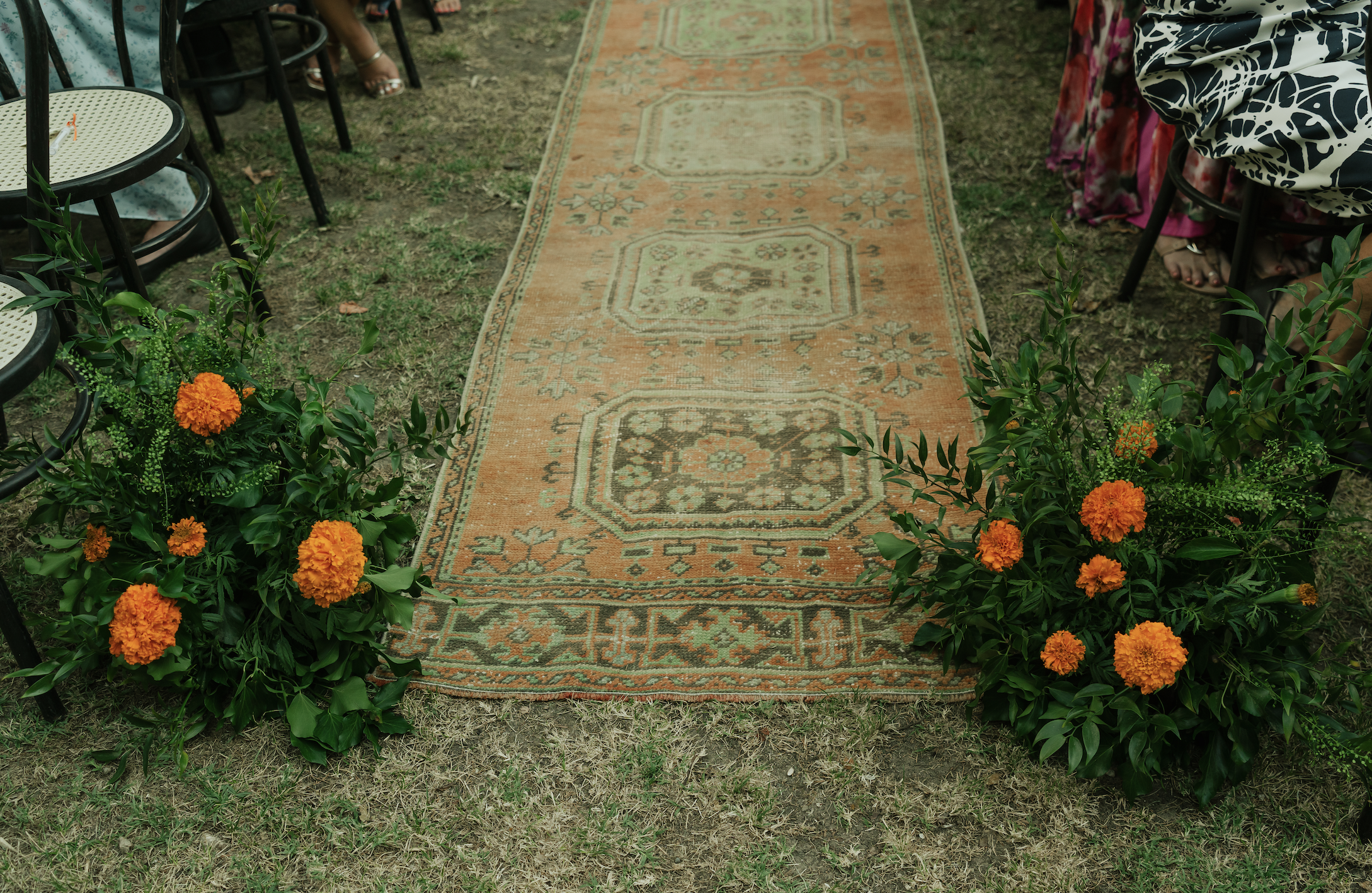 An outdoor wedding aisle decorated with orange flowers leading to chairs for guests, some of whom are partially visible.