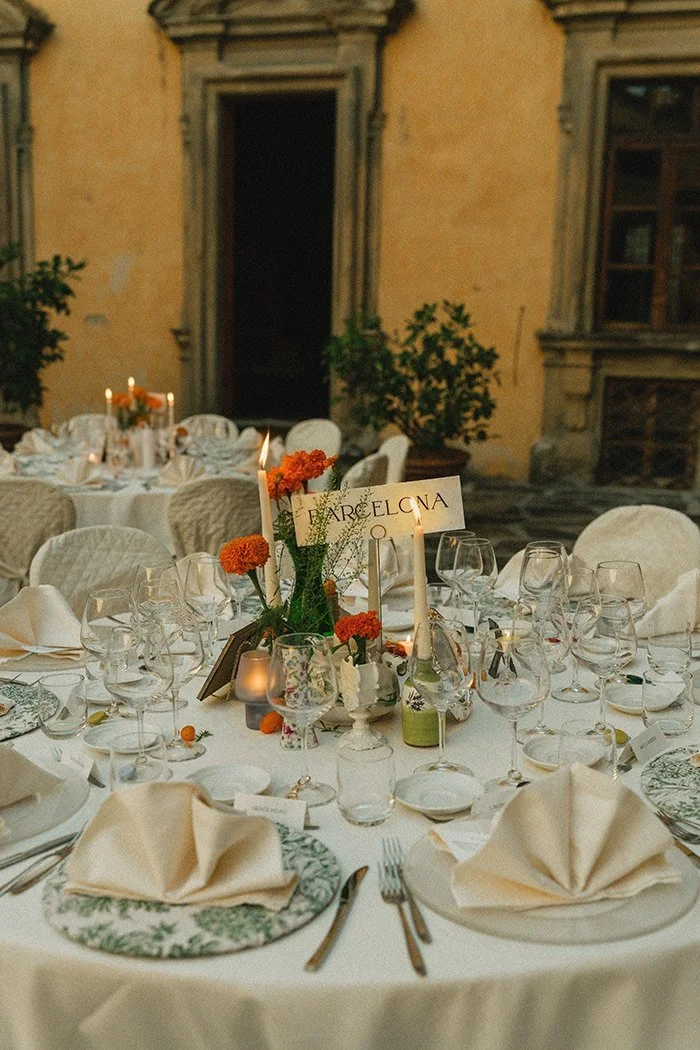 Elegant outdoor dining table in Italian castle courtyard set for a formal wedding event, surrounded by chairs with white covers, decorated with candles, orange marigold flowers, and vintage tableware, with an old building in the background.