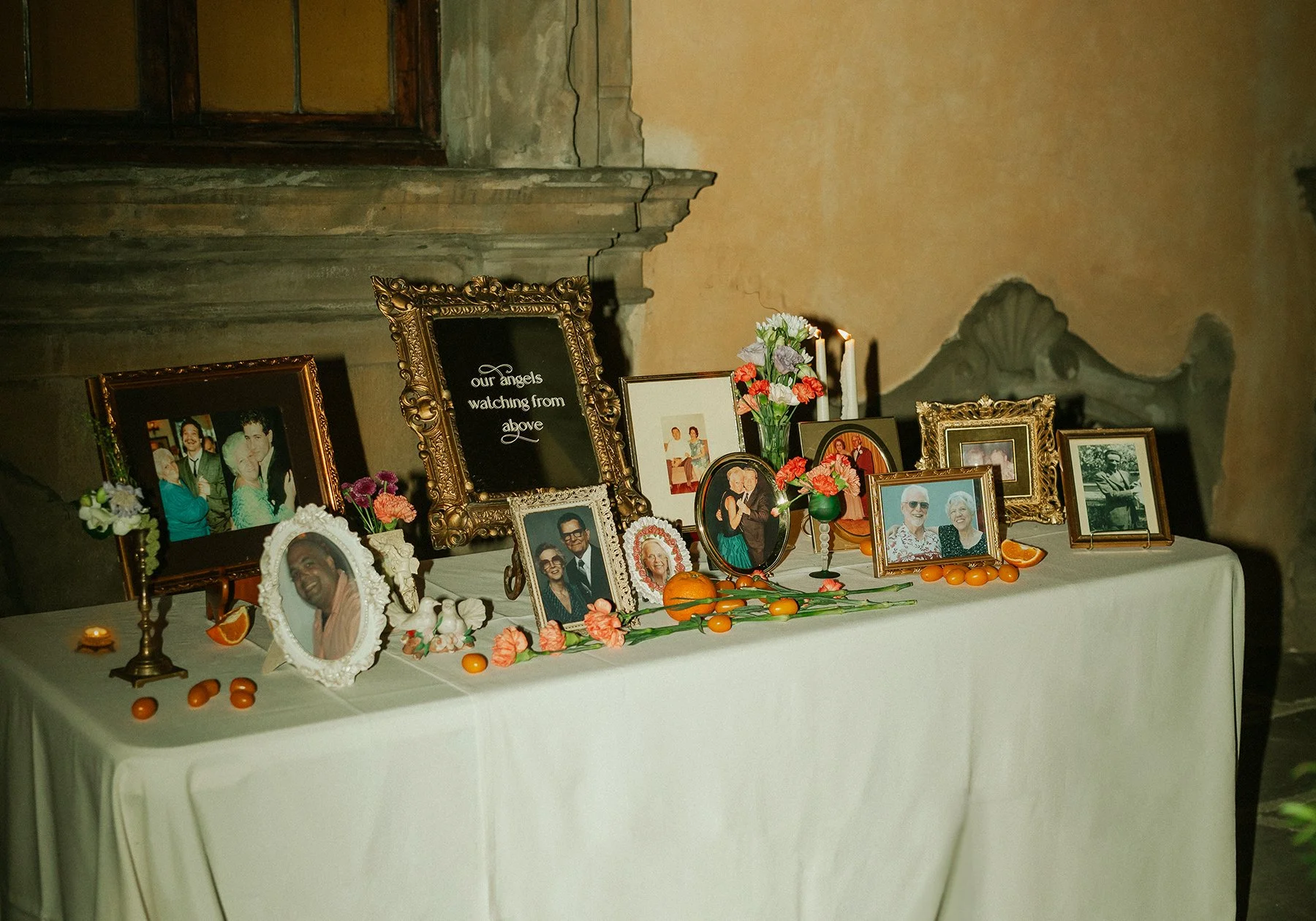 A memorial table with framed photos of deceased loved ones, orange flowers, candles, fruit, and a vintage mirrored sign that says 'our angels watching from above,' set against a rustic wall at a wedding.