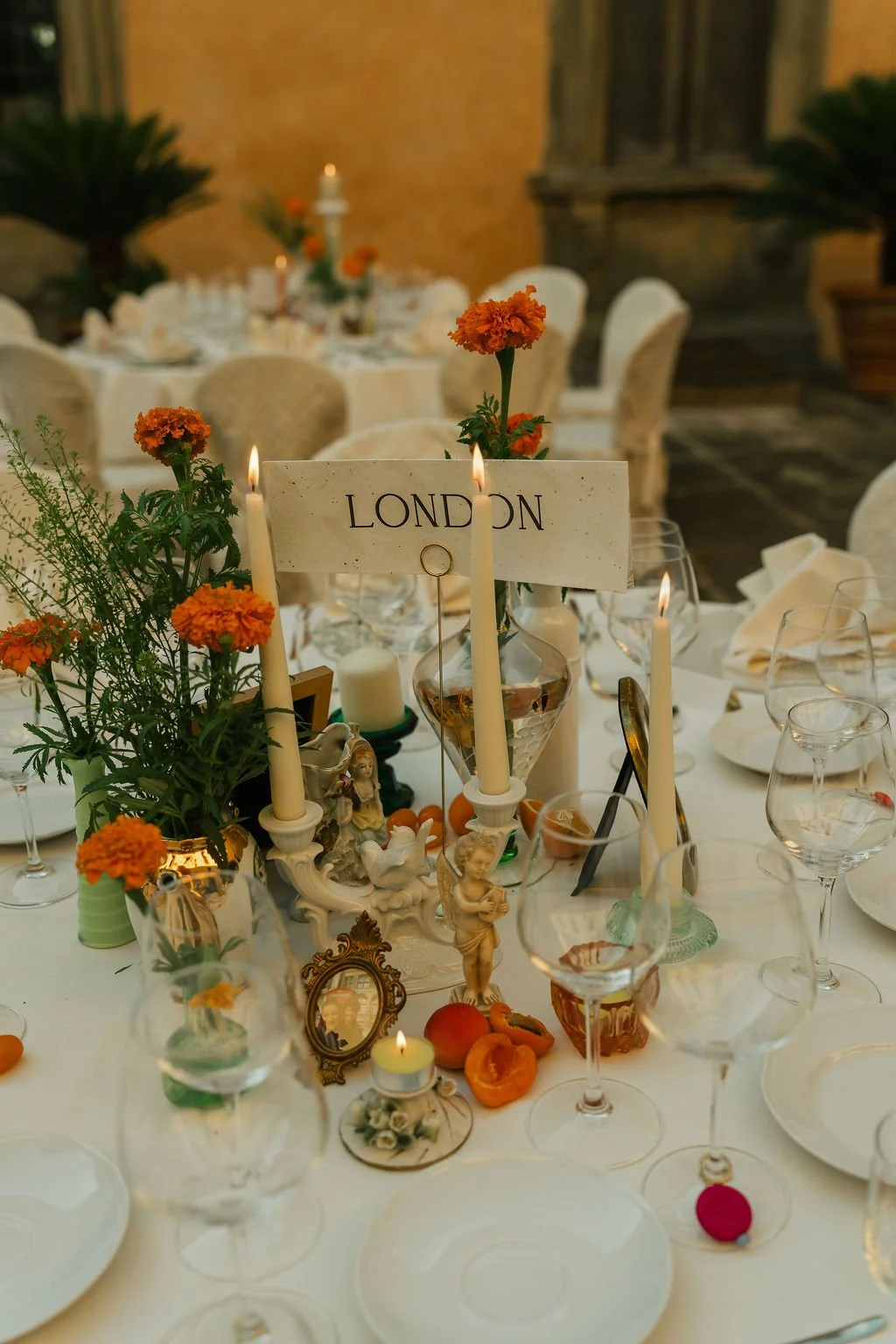 A decorated dining table centerpiece tablescape with orange flowers, candles, and figurines, labeled 'LONDON', set for a wedding dinner event in an elegant venue.