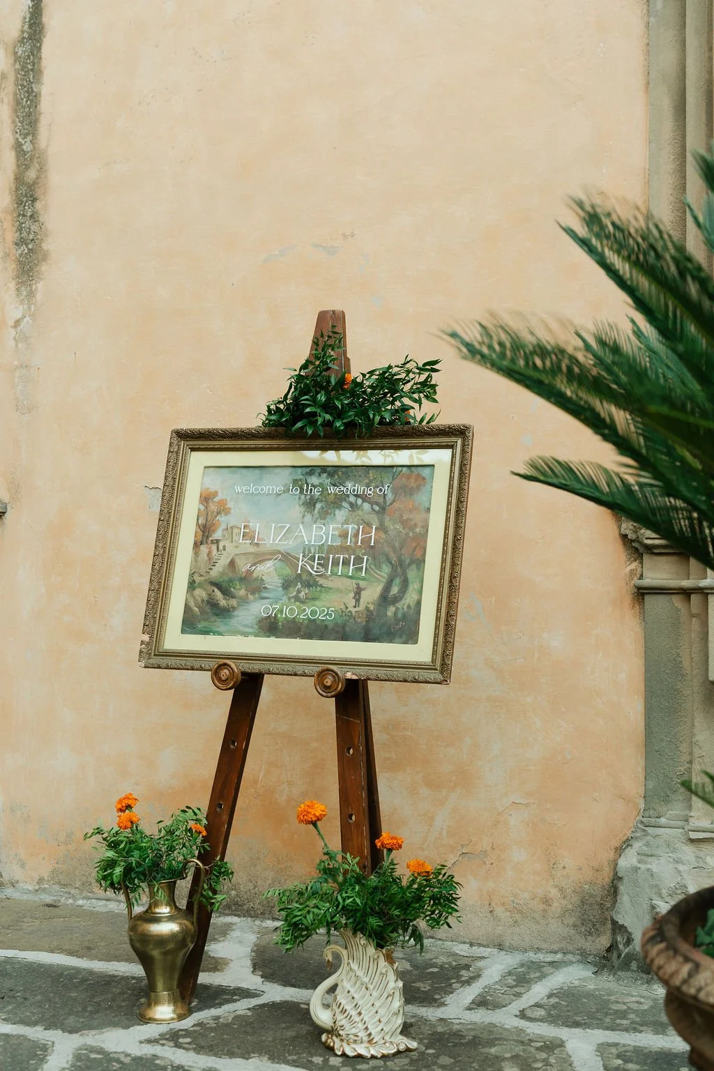 A vintage painting wedding welcome sign on an easel surrounded by potted plants, greenery, and orange flowers, with a textured beige wall in the background.