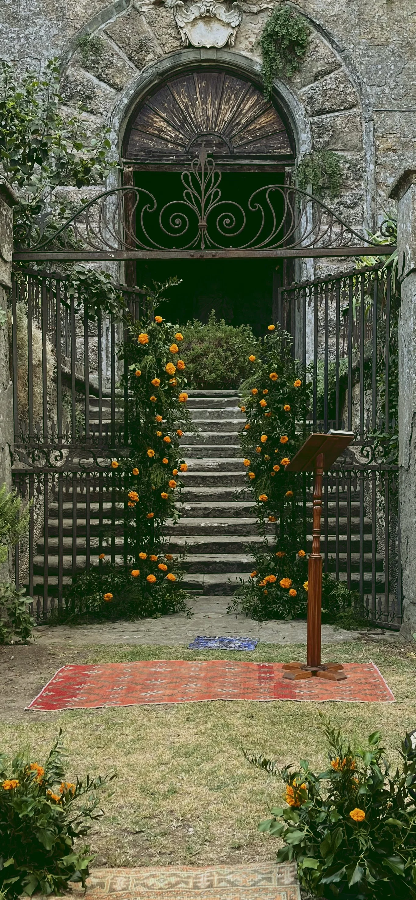 Stone staircase adorned with orange marigold flower arch at wedding leads to an arched door with bronze detailing set in a stone wall, surrounded by greenery and iron gate, with a small vintage Persian rug and wooden stand on grassy ground.