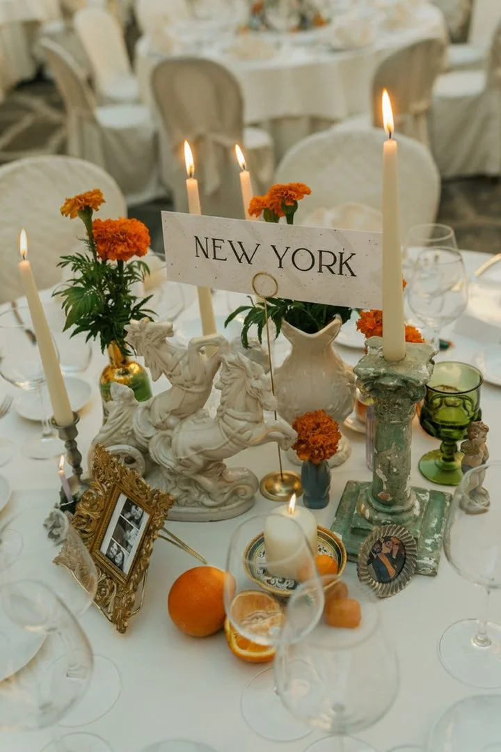 A decorated table at a wedding with a centerpiece featuring a "New York" sign, orange flowers, white candles, and antique framed photographs, set in an elegant setting with multiple vintage glassware and white tablecloths.