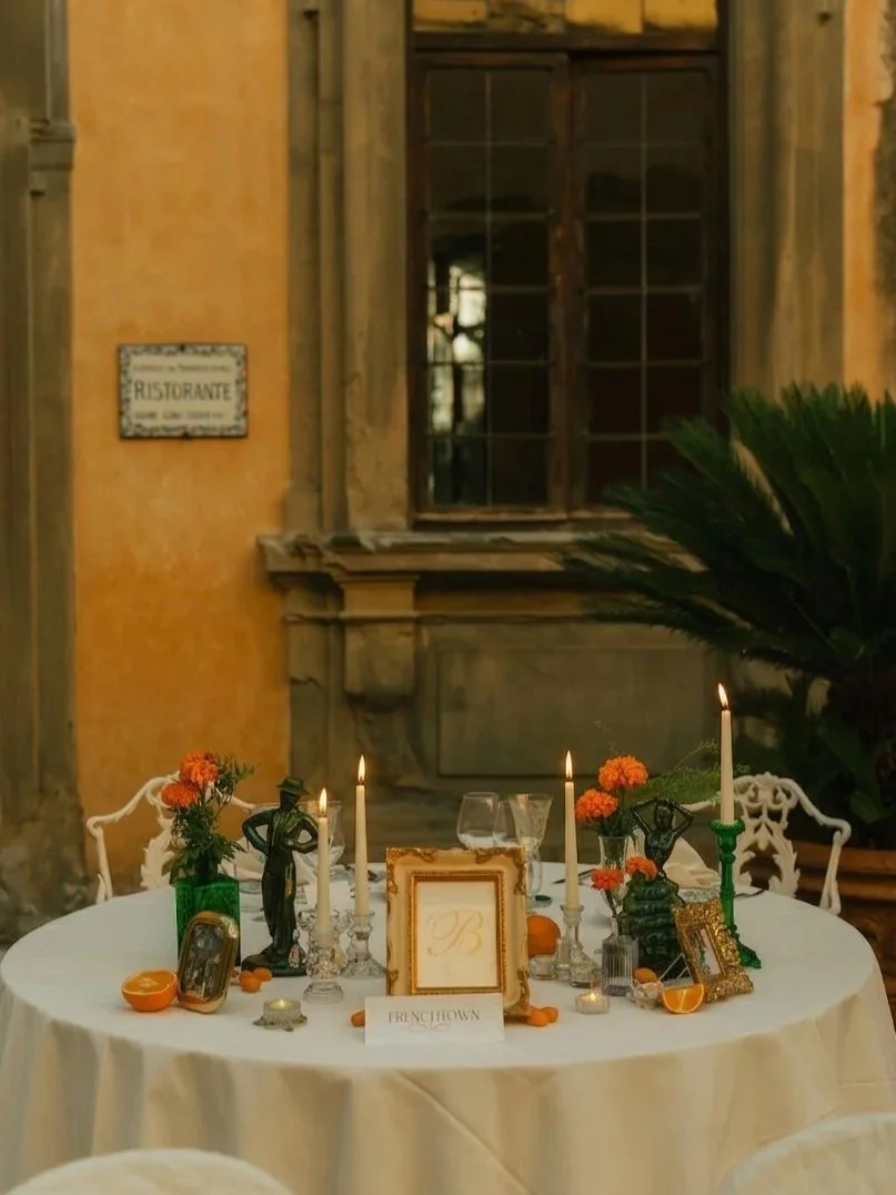 A decorated sweetheart wedding round table with white tablecloth, candles, orange slices, and floral arrangements in a dimly lit setting with an old building facade in the background, indicating a festive or elegant wedding event.