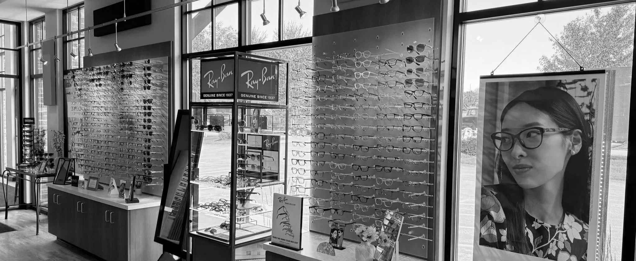 Inside an eyewear store with sunglasses and glasses displayed on stands and racks, Ray-Ban advertisements, and a large photograph of a woman wearing glasses near the window.