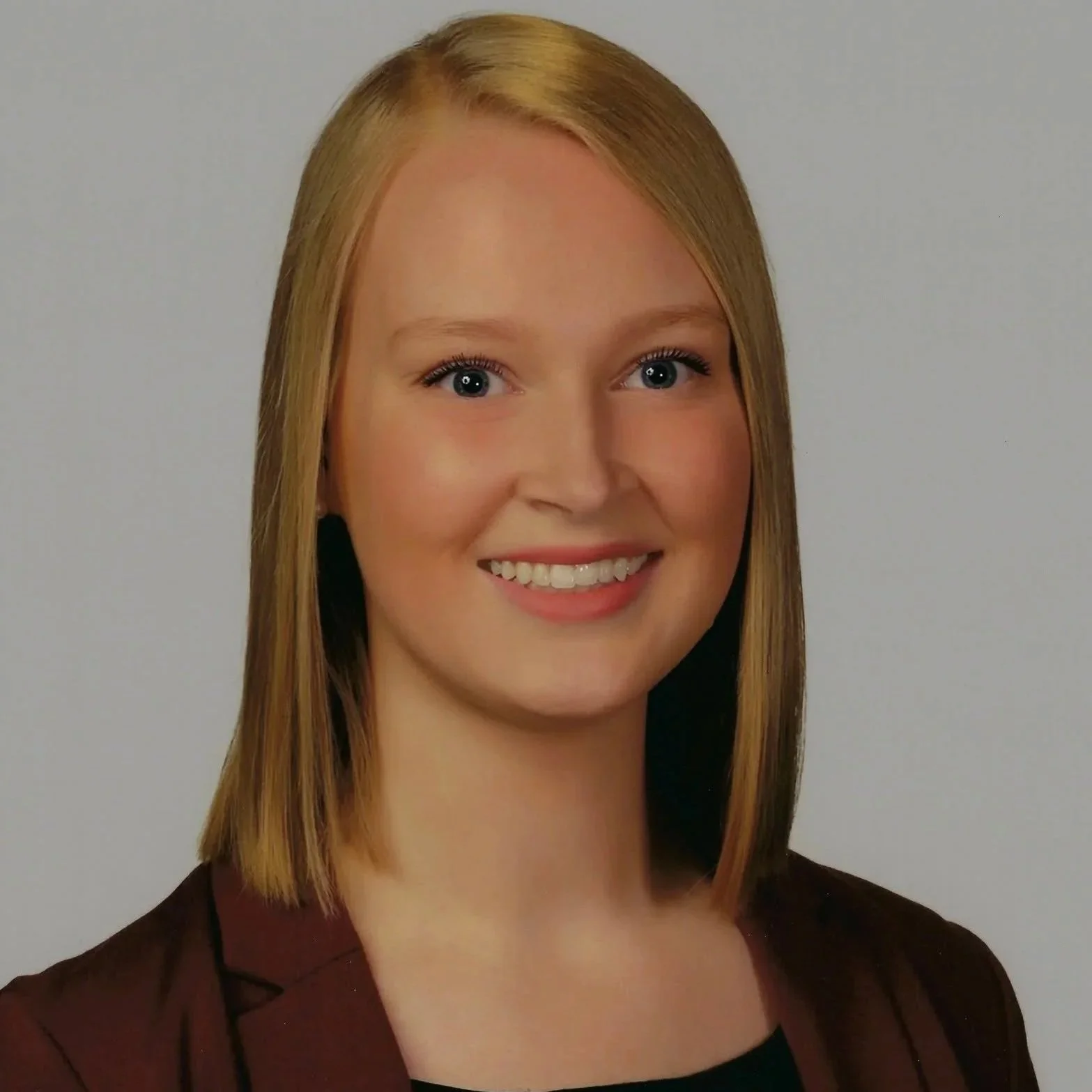 Headshot of a woman with shoulder-length blond hair, smiling, wearing a dark blazer over a black top, against a plain gray background.