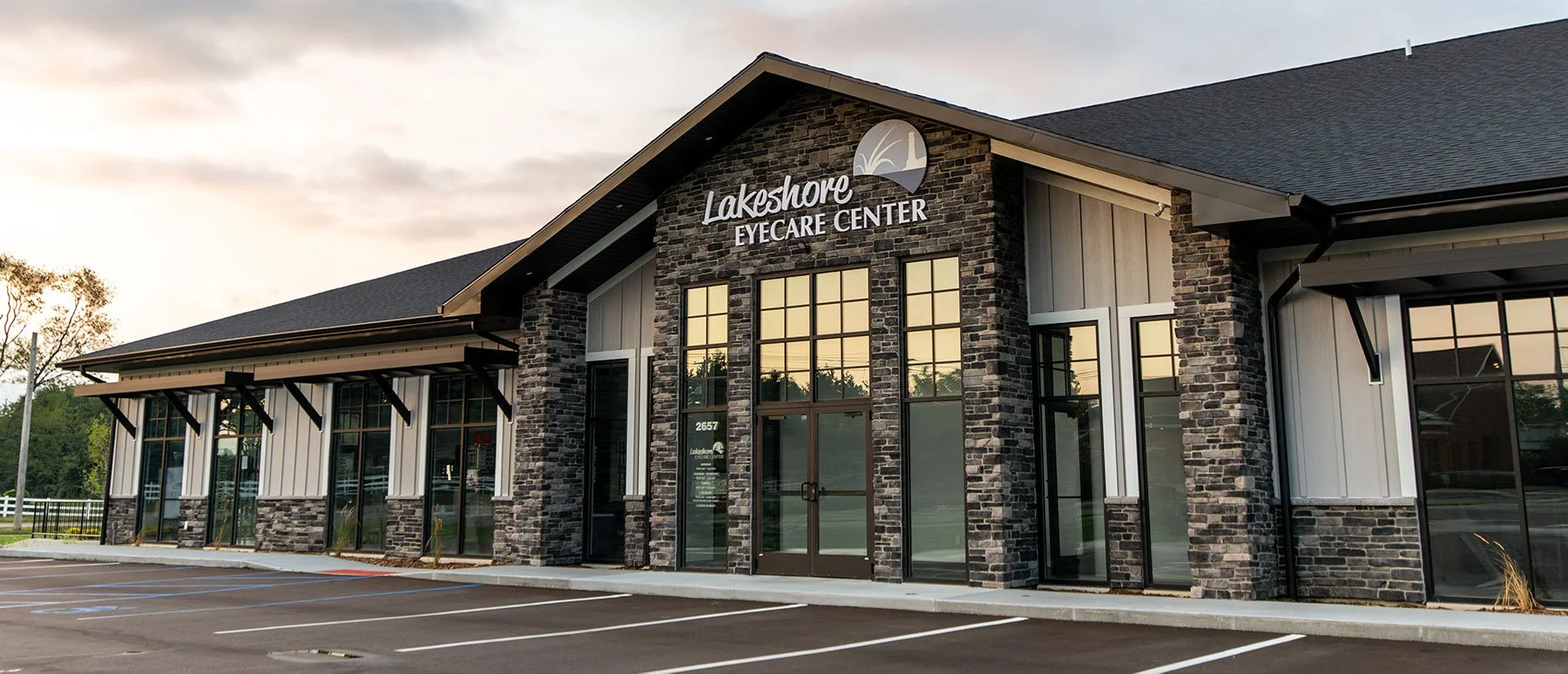 Exterior of Lakeshore Eyecare Center building with stone and wood facade, large glass windows, and parking lot in foreground at sunset.