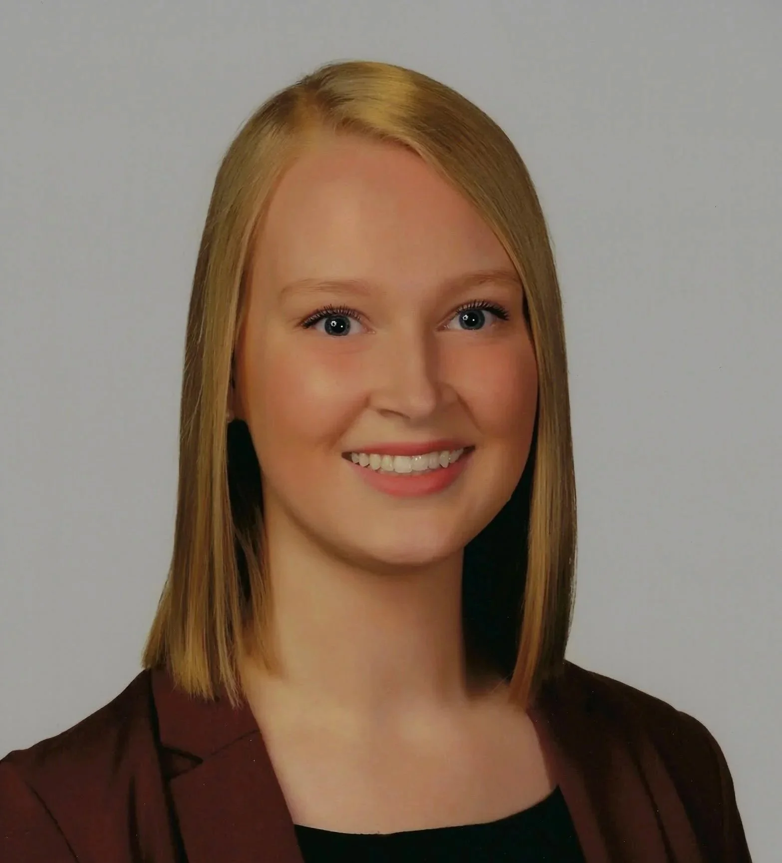Portrait of a woman with straight blonde hair, blue eyes, smiling, wearing a black top and a dark brown blazer.