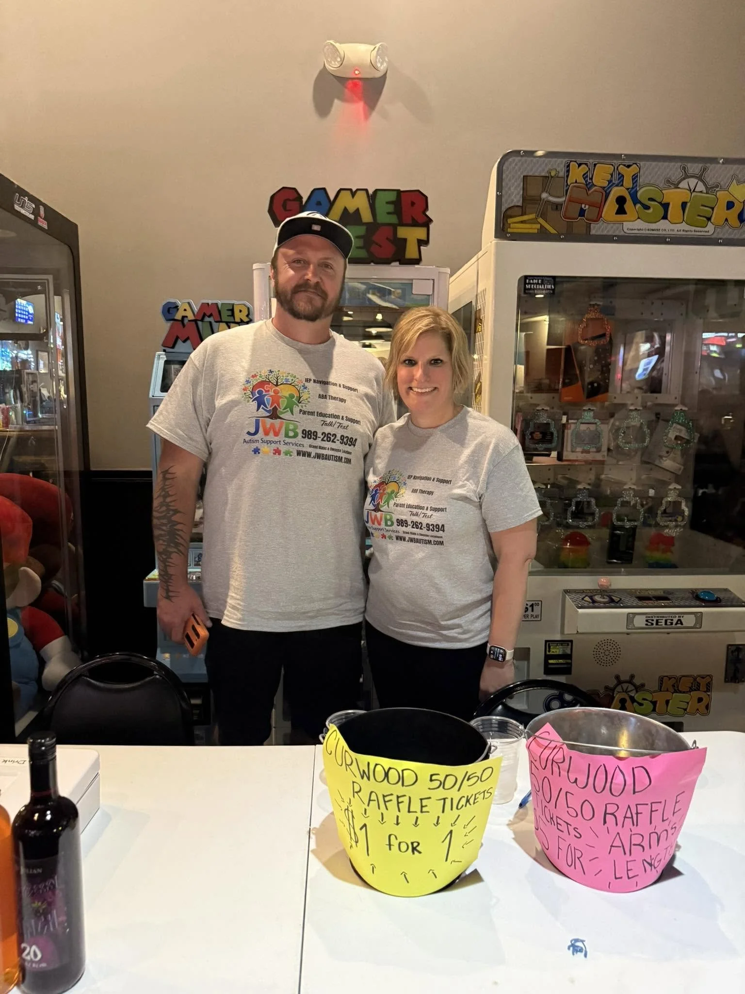 A man and woman standing behind a table at a raffle event, with signs advertising rainbow raffles, tickets, and prices, inside an arcade or game area with claw machines and arcade games in the background.