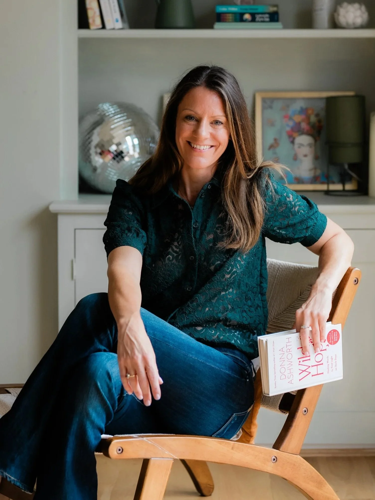 Smiling woman in a teal blouse sitting in a wooden chair and holding a copy of “Wild and Worthy” at home.