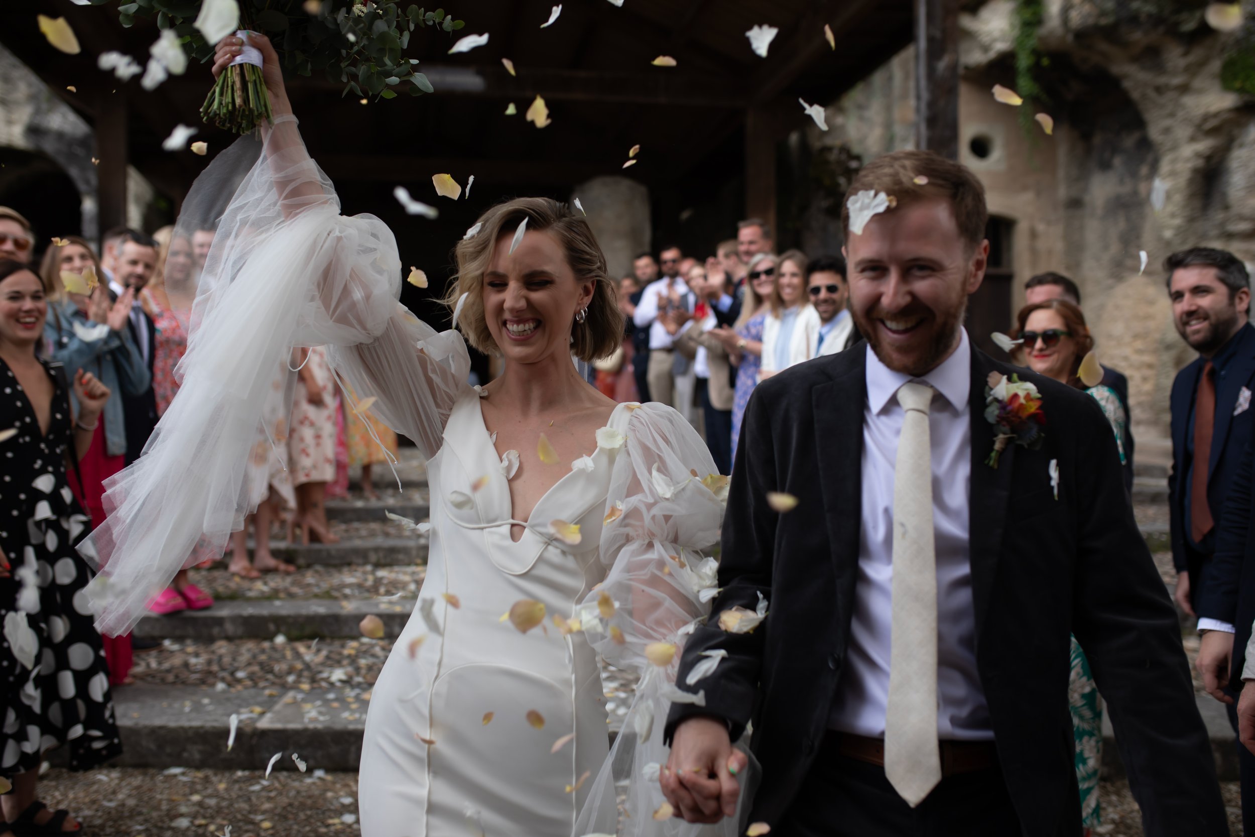A newlywed couple walking hand in hand at their wedding celebration, surrounded by friends and family, with confetti falling in the air and a rustic outdoor setting in the background.