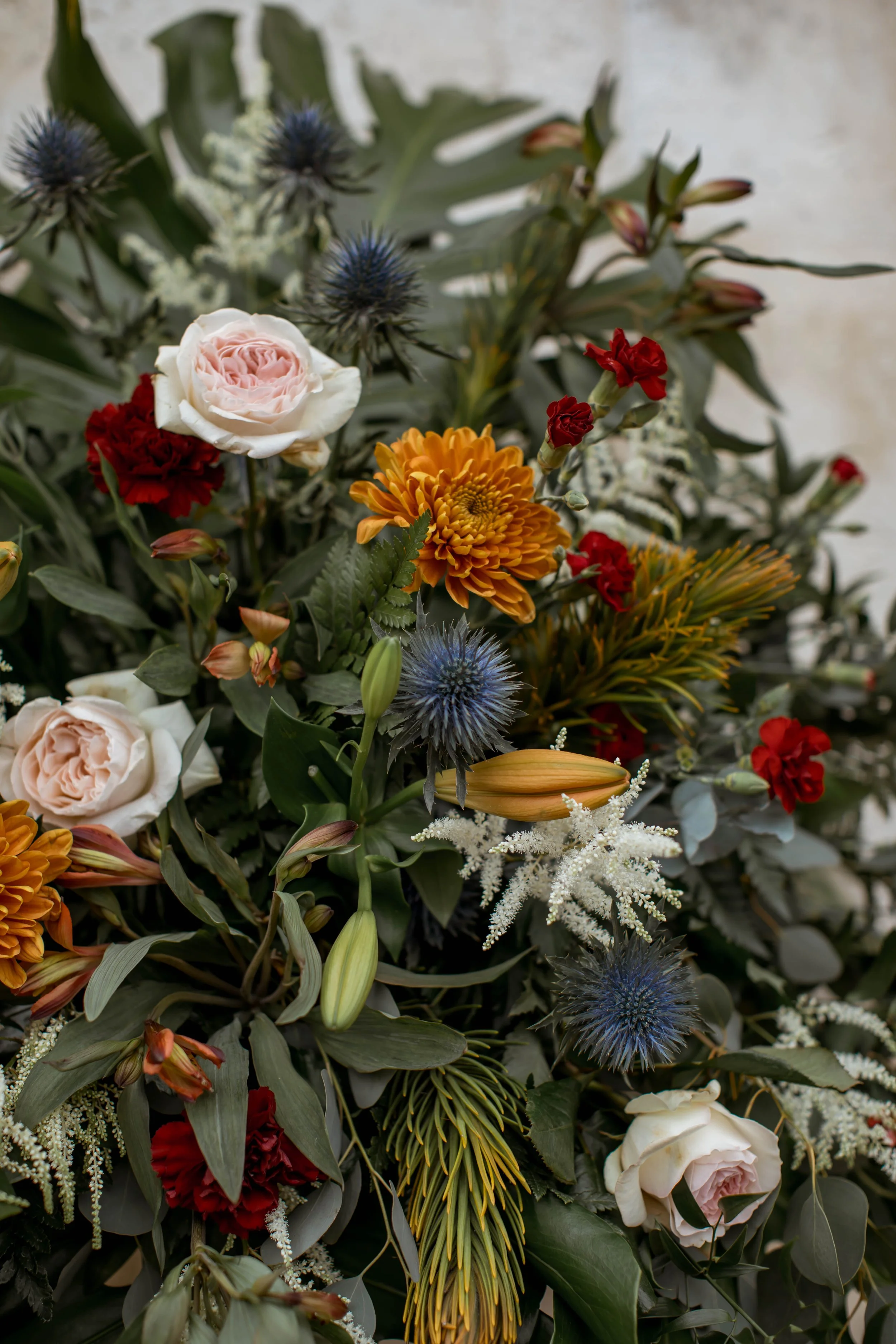 A colorful arrangement of various flowers, including pink roses, red carnations, yellow chrysanthemums, white astilbe, and blue thistles, with green foliage.