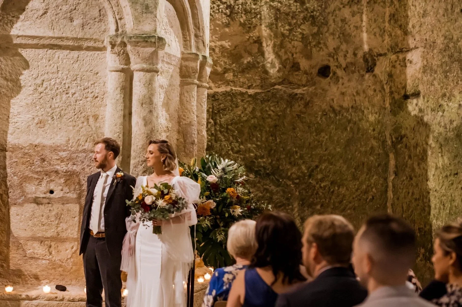 A bride and groom standing in front of a stone wall during their wedding, with guests seated in front of them. The bride holds a bouquet and wears a white dress, while the groom wears a suit.