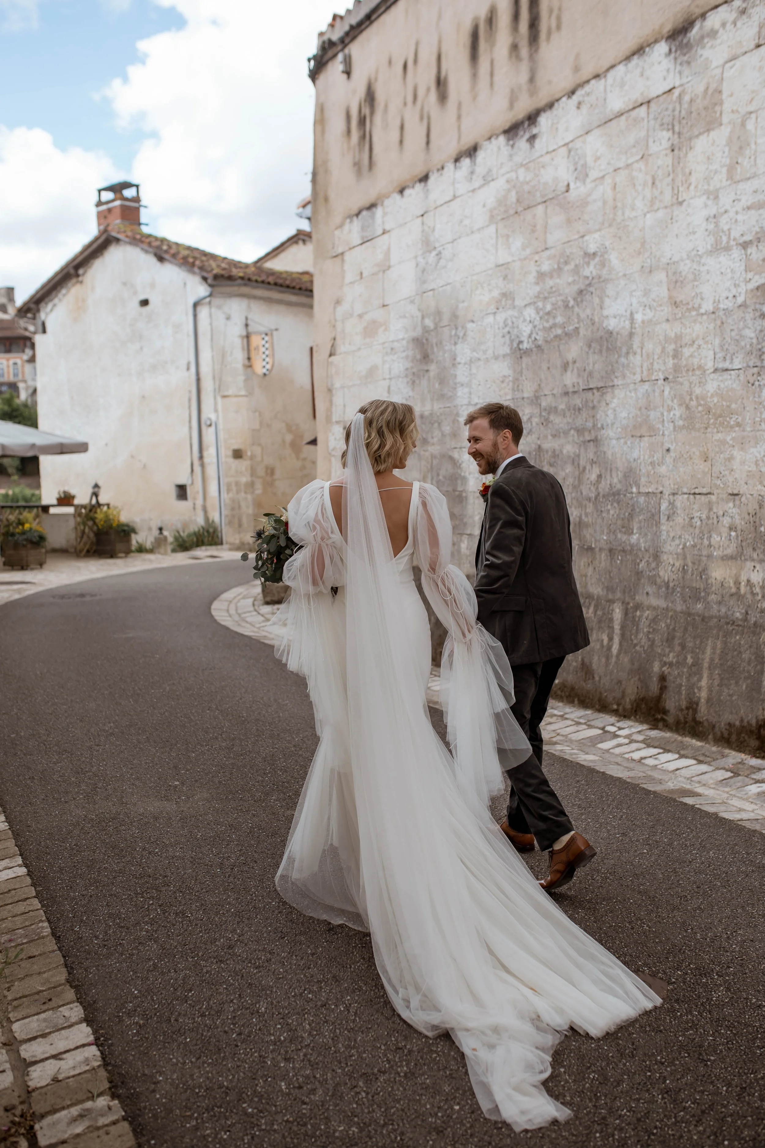 A bride and groom walk together in a quaint outdoor setting with old stone buildings. The bride wears a long white gown with flowing tulle, and the groom wears a dark suit and brown shoes. They smile at each other, holding hands.