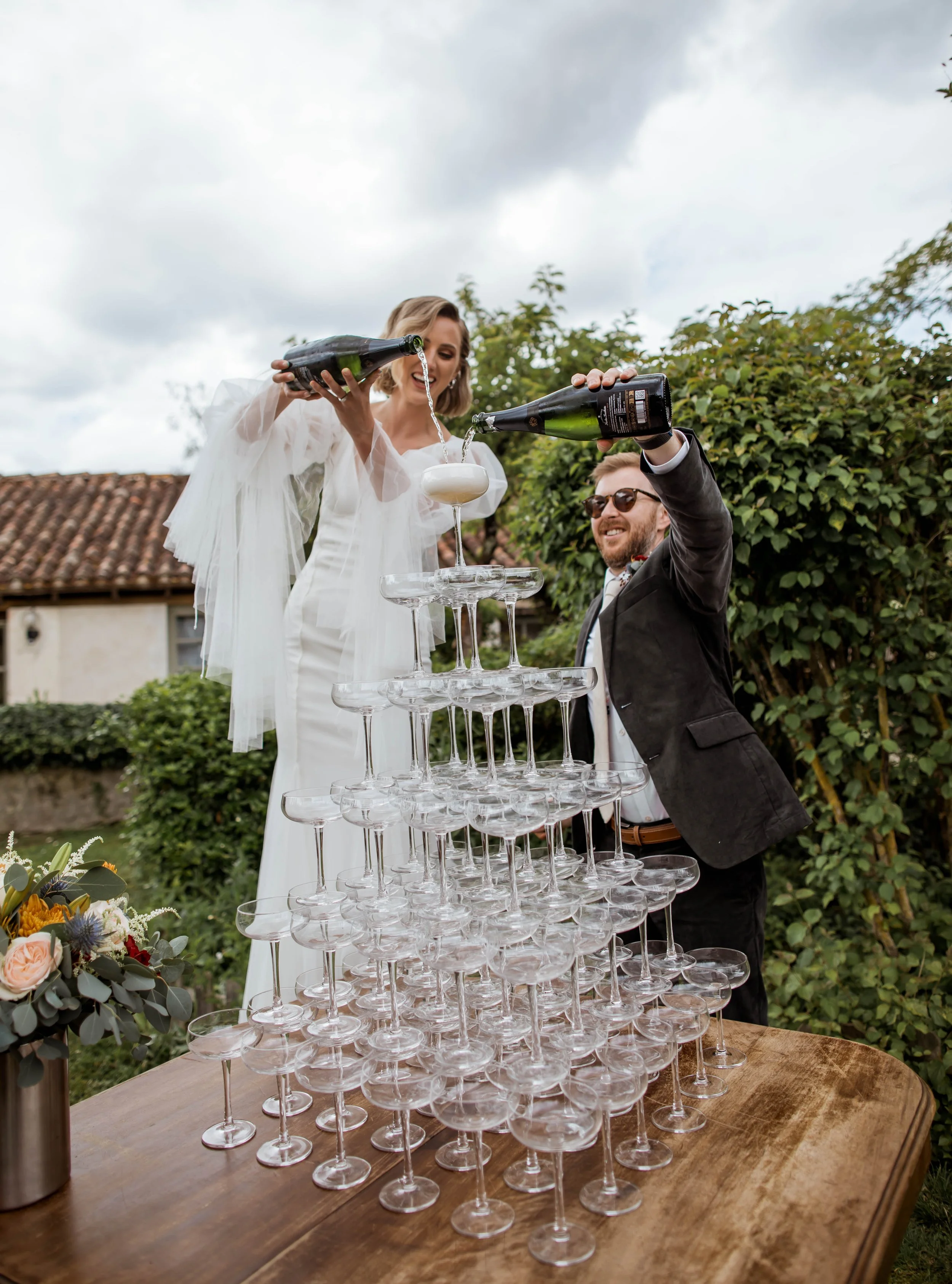 A woman in a white dress and a man in a black tuxedo are celebrating outdoors, pouring champagne into a champagne tower made of stacked glassware. The scene takes place in a garden with lush greenery, a building with a tiled roof, and a cloudy sky in the background.