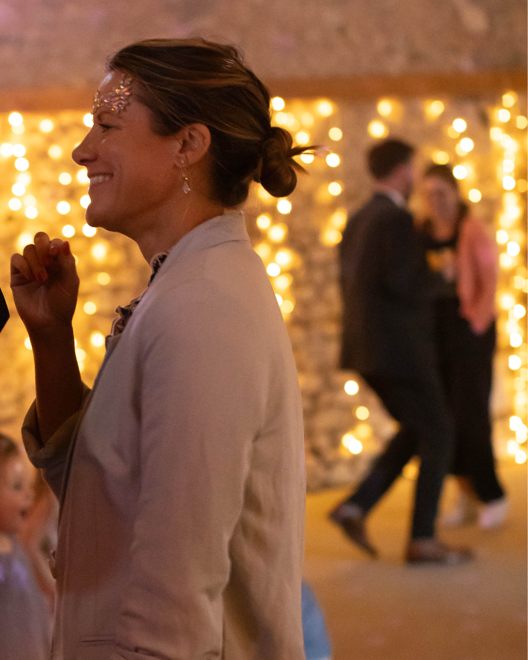 A woman smiling with glitter on her forehead at a party or celebration, with blurred people dancing and string lights in the background.
