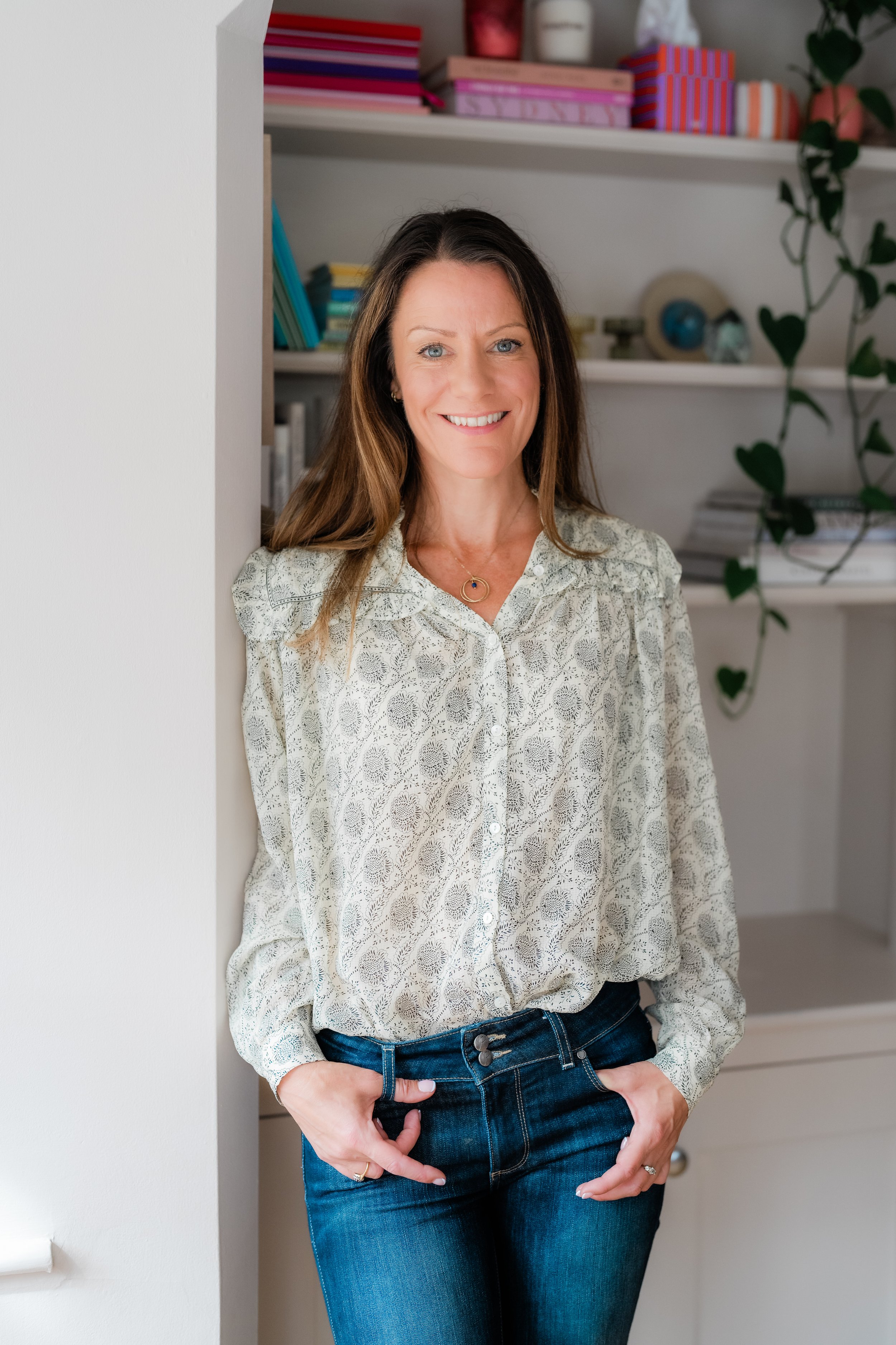 Smiling woman in a floral blouse and blue jeans standing in front of bookshelves and houseplants
