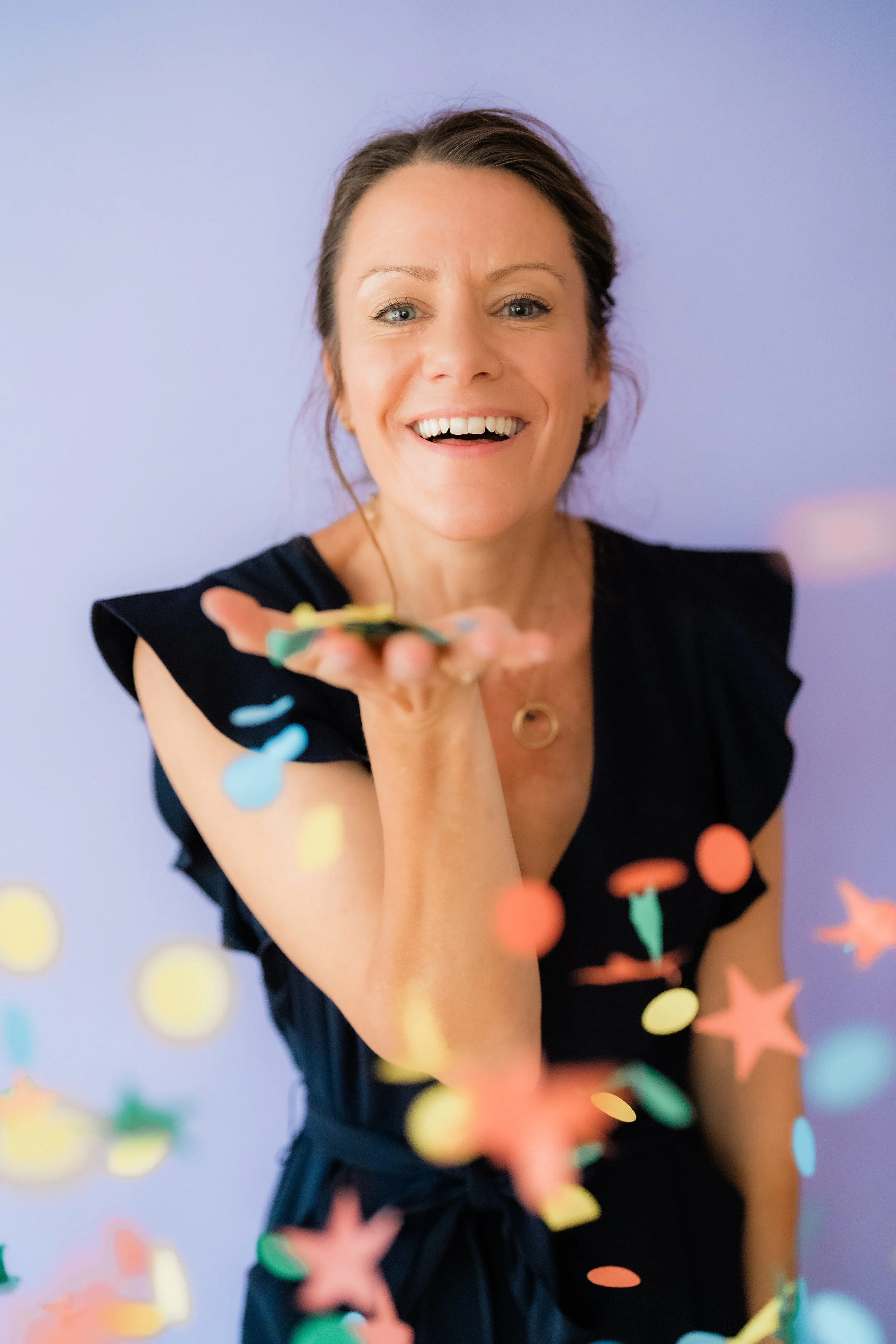 Smiling woman in a black shirt holding colorful confetti against a lavender background
