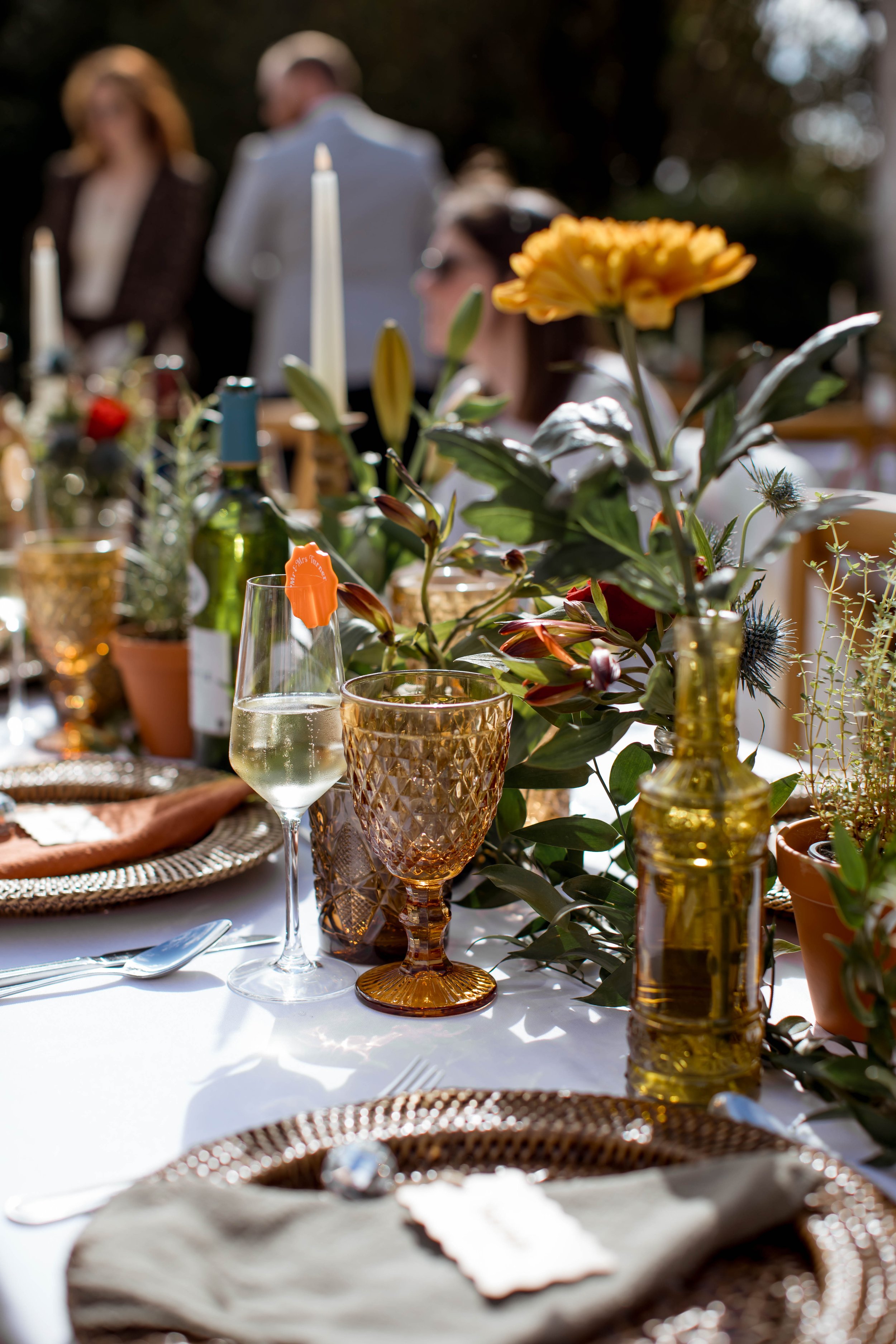 Close-up of a table setting with a floral centerpiece, glasses of wine, and gold-colored decor, with blurred guests in the background at an outdoor event.