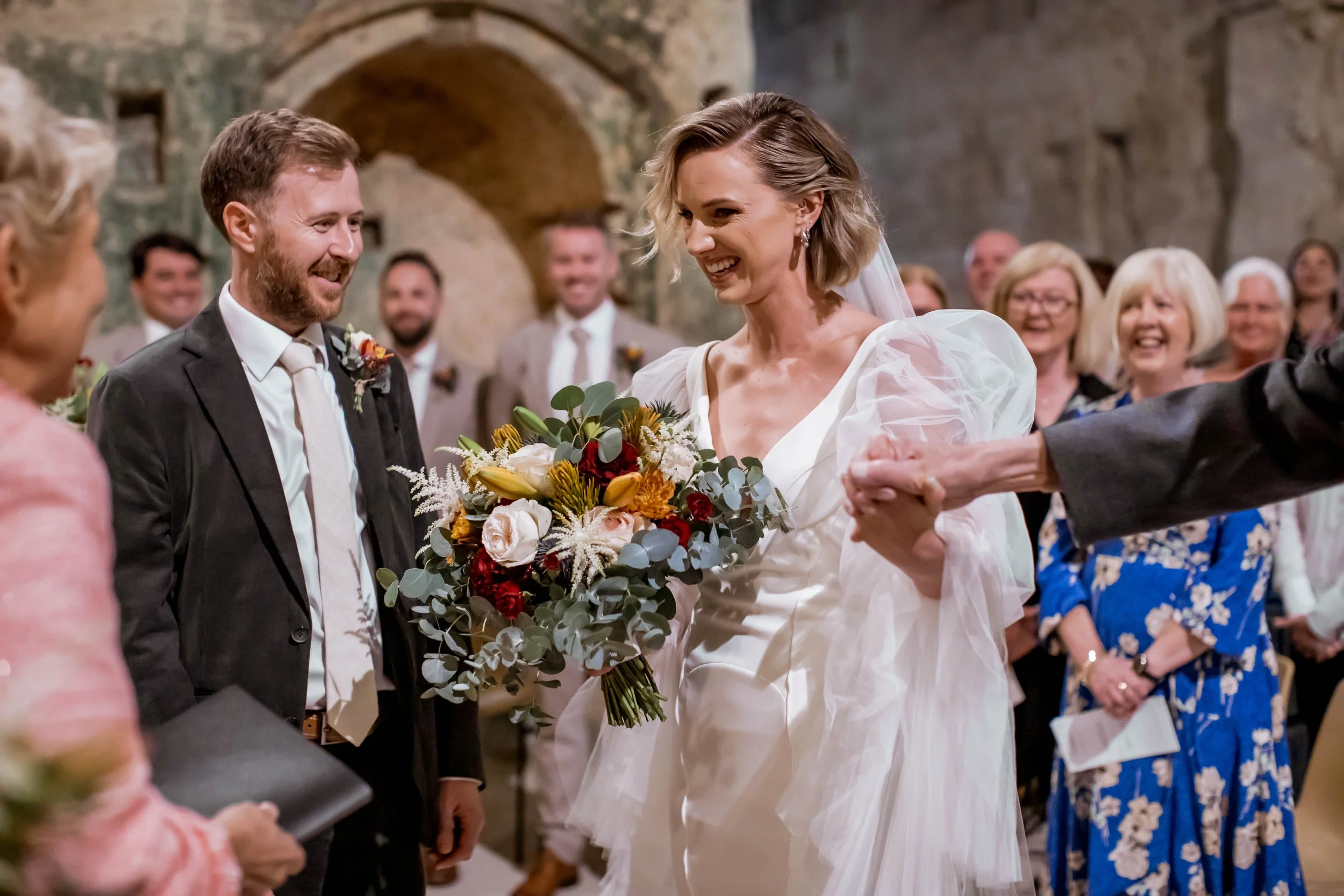 A bride and groom holding hands during their wedding ceremony, smiling at each other, with guests in the background in an indoor venue with stone walls.