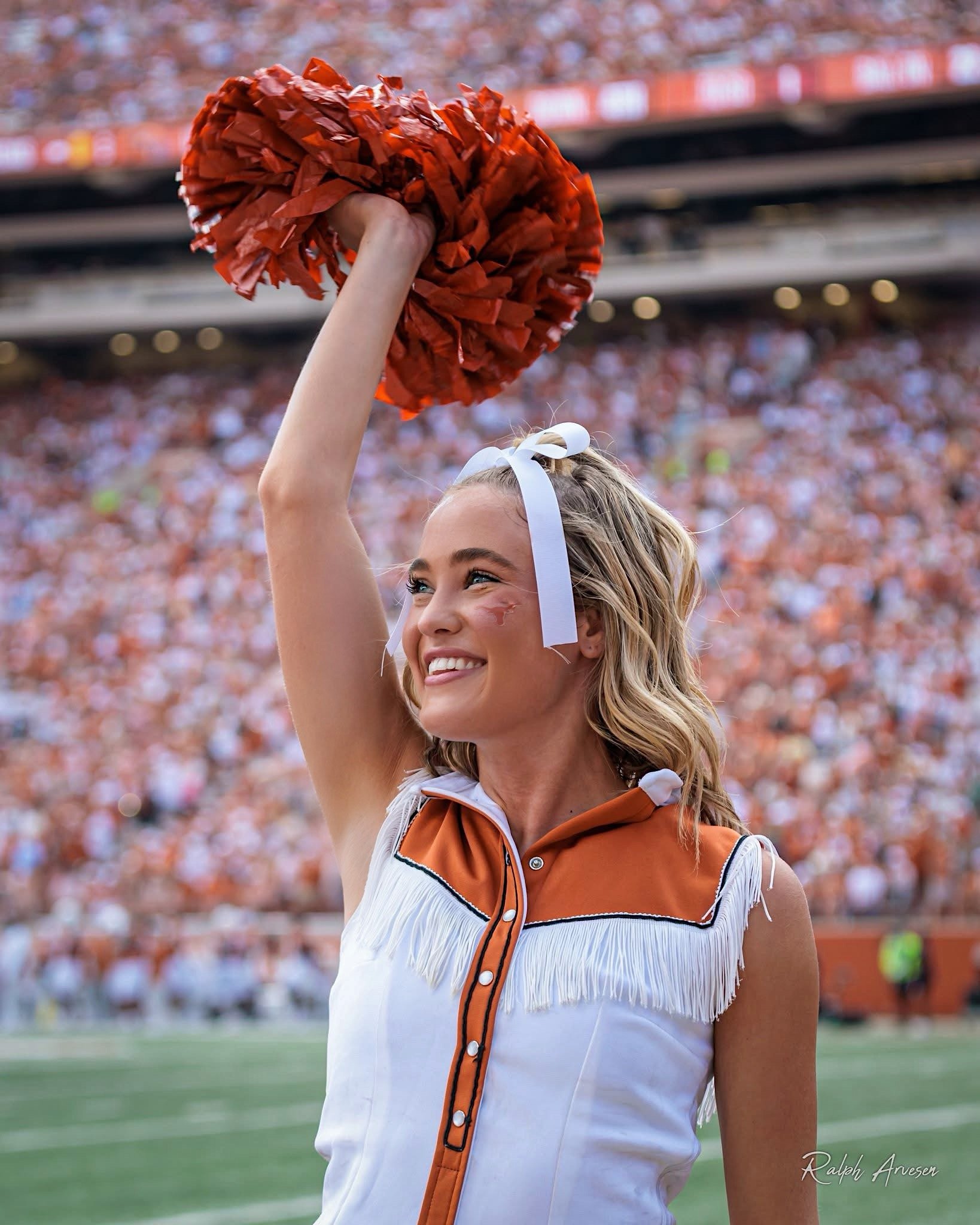 Close-up of a cheerleader in a white and orange cheerleading uniform holding an orange pom-poofer on a sports field with a large crowd in the background.
