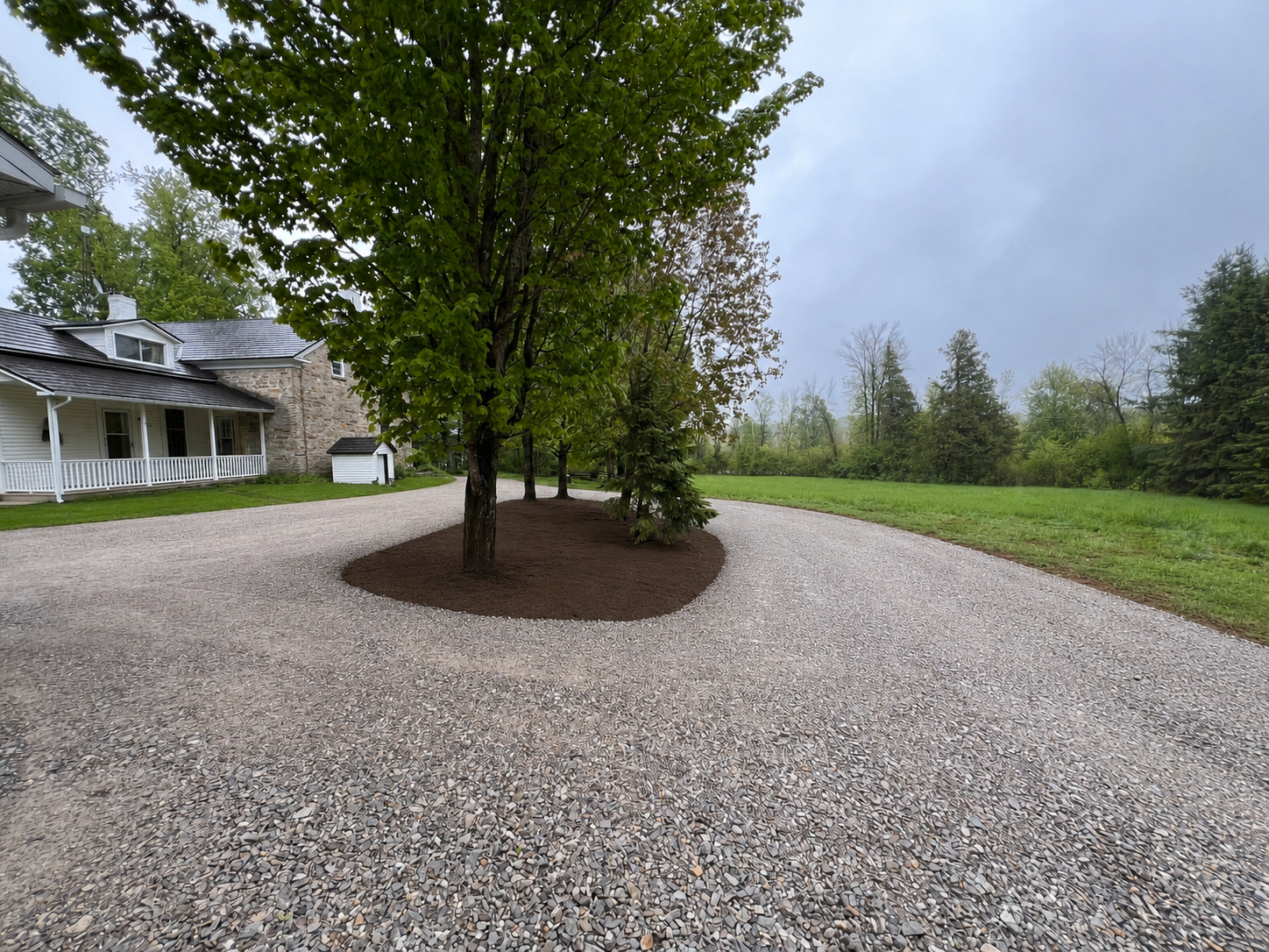 A gravel driveway curves around a landscaped area with trees and grass, with a house in the background.