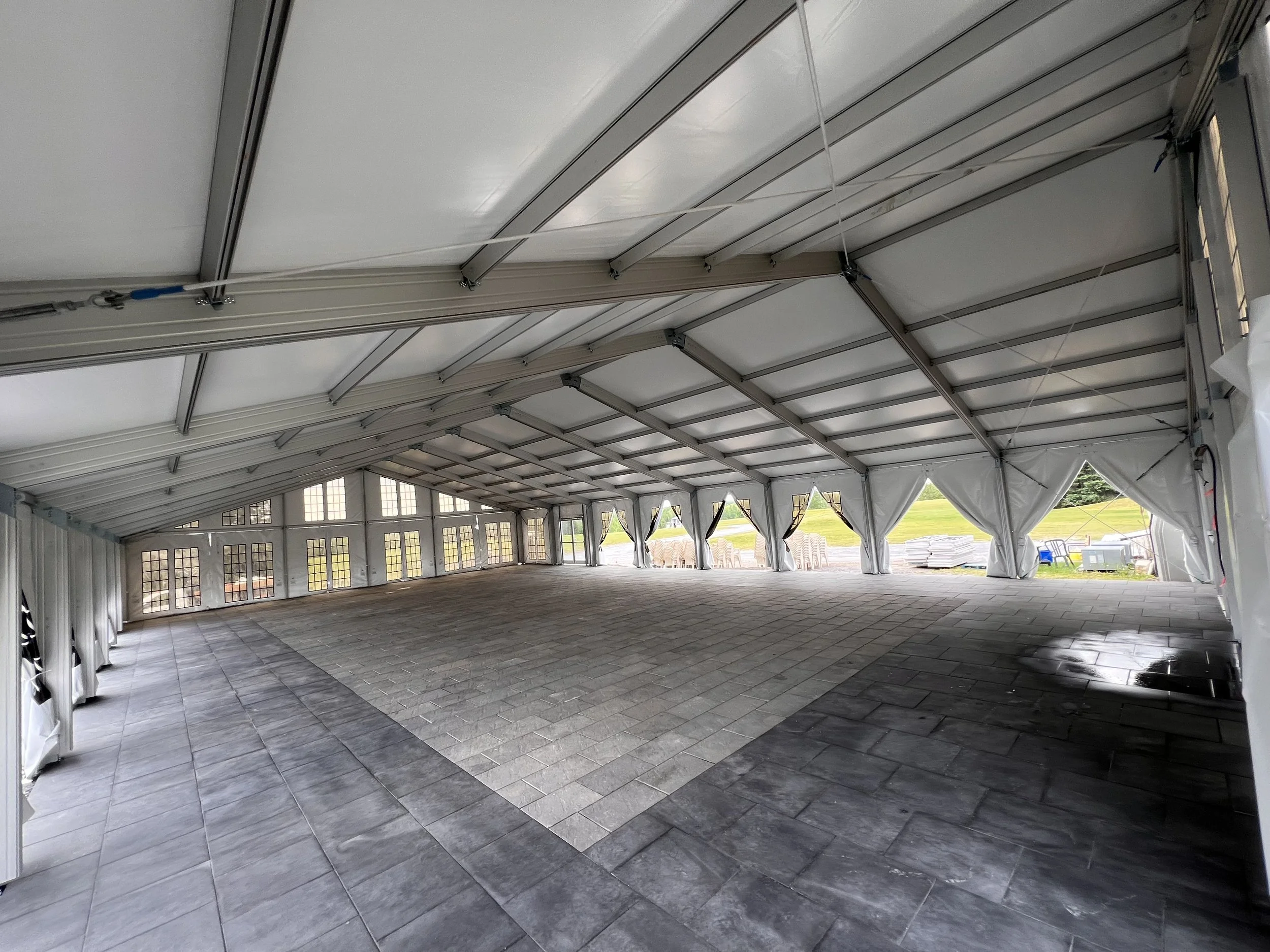 The interior of a large tent with a white ceiling, gray metal support beams, and dark gray tiled flooring. The sides of the tent have windows and open flaps revealing chairs, tables, and a grassy outdoor area beyond.