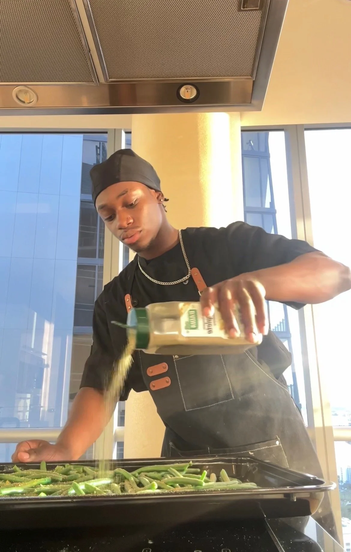 A young man in a black apron and chef's hat adds seasoning to a pan of green beans in a modern kitchen with large windows.