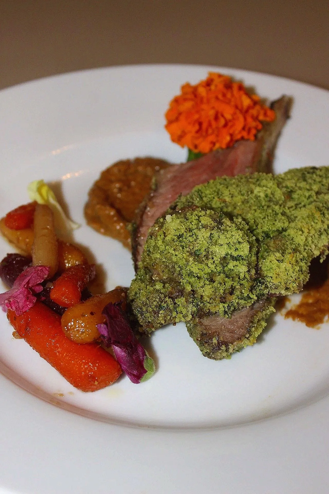 Close-up of a white plate with roasted meat topped with green herb crust, likely surrounded by vegetables and garnishes.