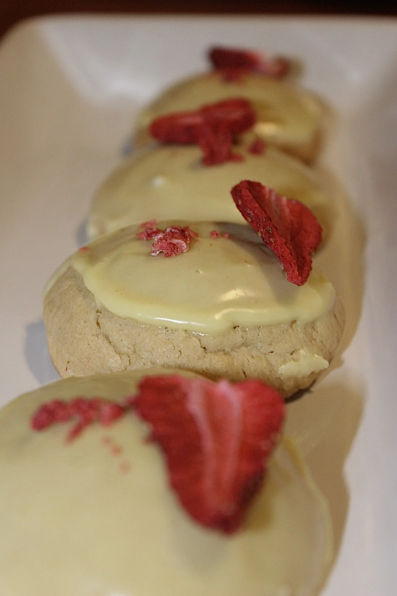Close-up image of small cookies with white chocolate coating and red dried fruit pieces on top, arranged in a row.