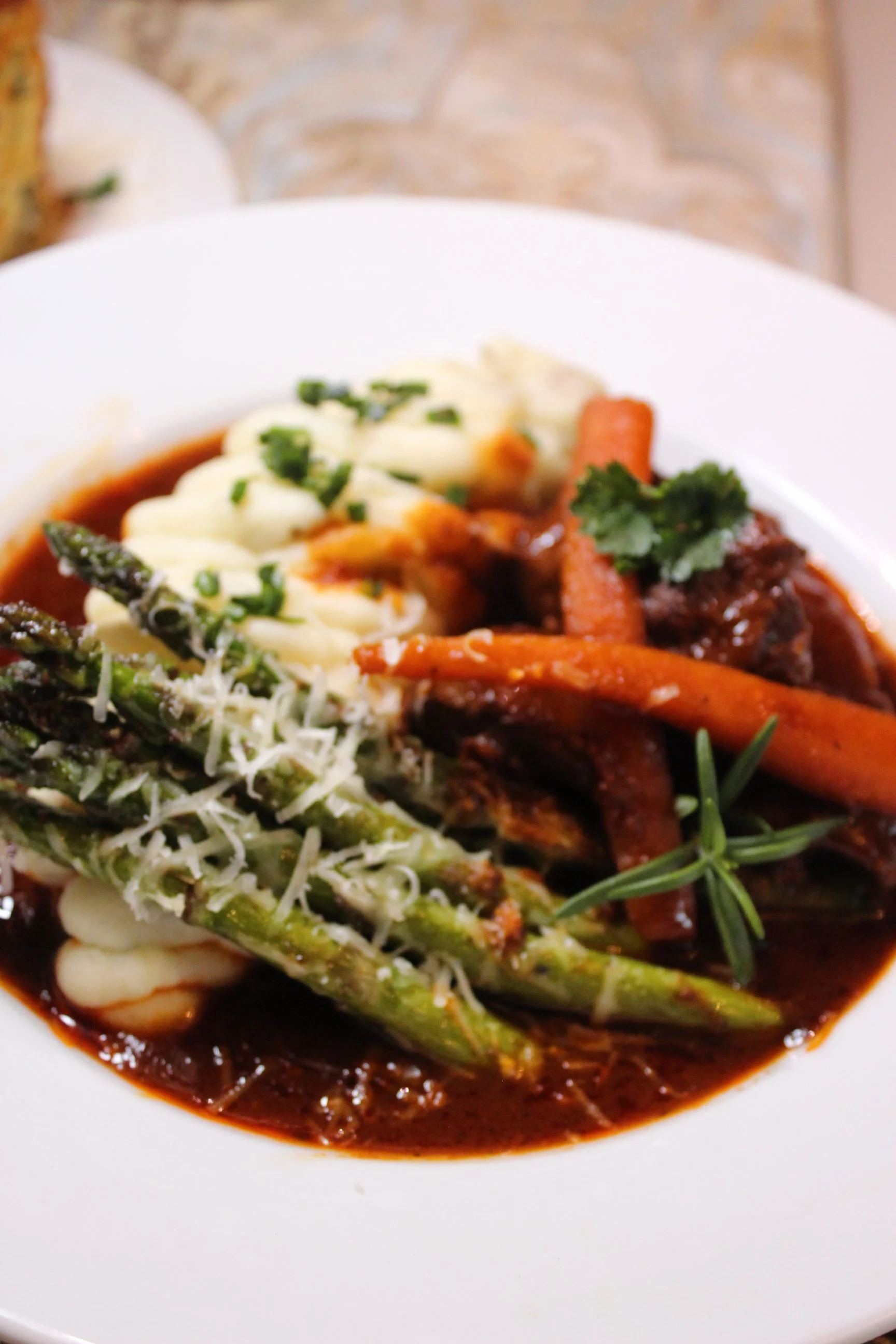 A bowl of beef stew with carrots and herbs, topped with mashed potatoes and green chives, served with green beans and shredded cheese on top.
