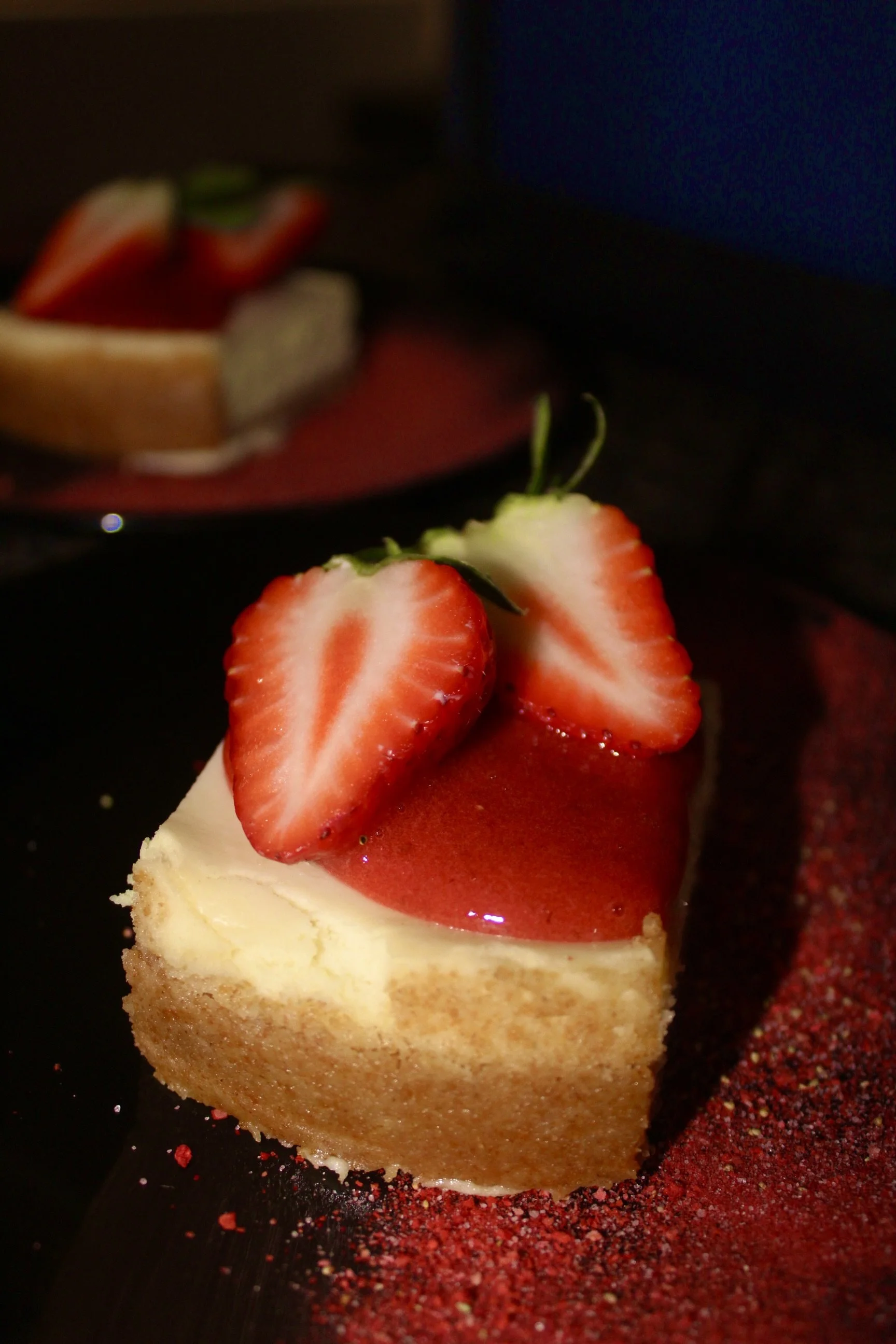 A slice of cheesecake topped with strawberries and strawberry glaze, garnished with green strawberry leaves, on a black plate with red seasoning around it in the foreground, with a blurred background featuring additional strawberries and a dessert.