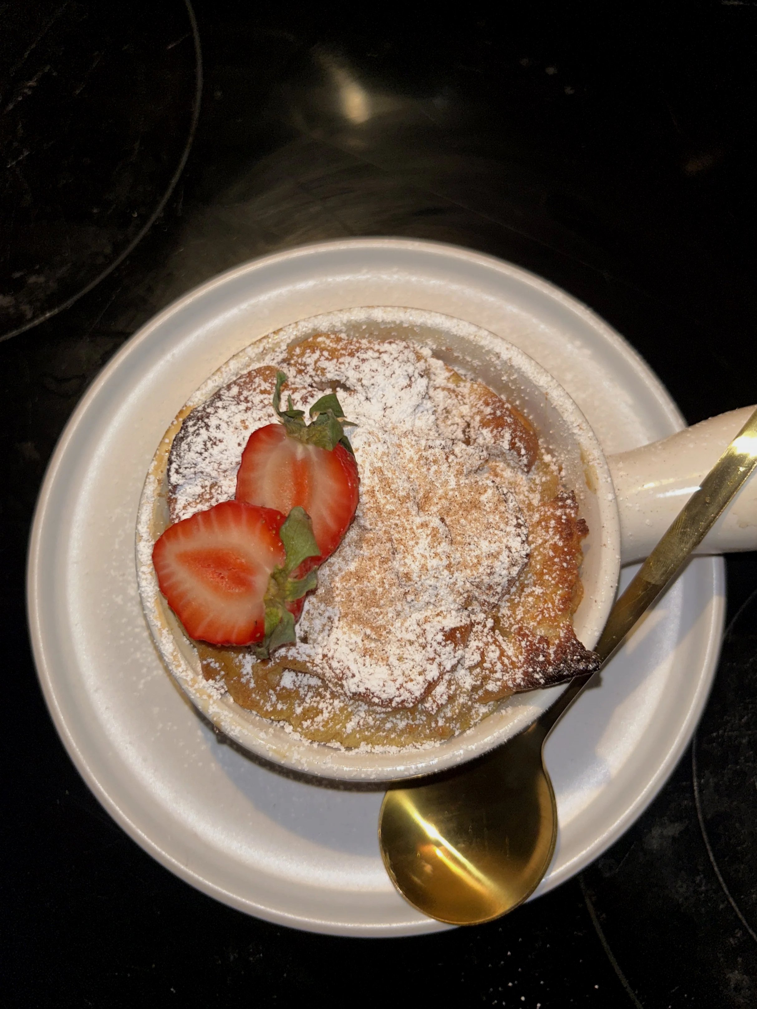 A bowl of caramel custard topped with powdered sugar and sliced strawberries with a gold spoon.