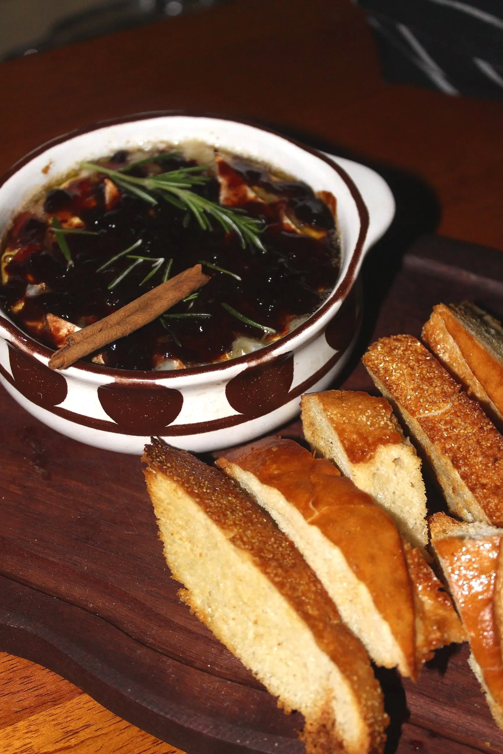 A ceramic dish of Chinese black bean stew garnished with a sprig of rosemary and a cinnamon stick, accompanied by slices of fried bread or cake on a wooden tray.