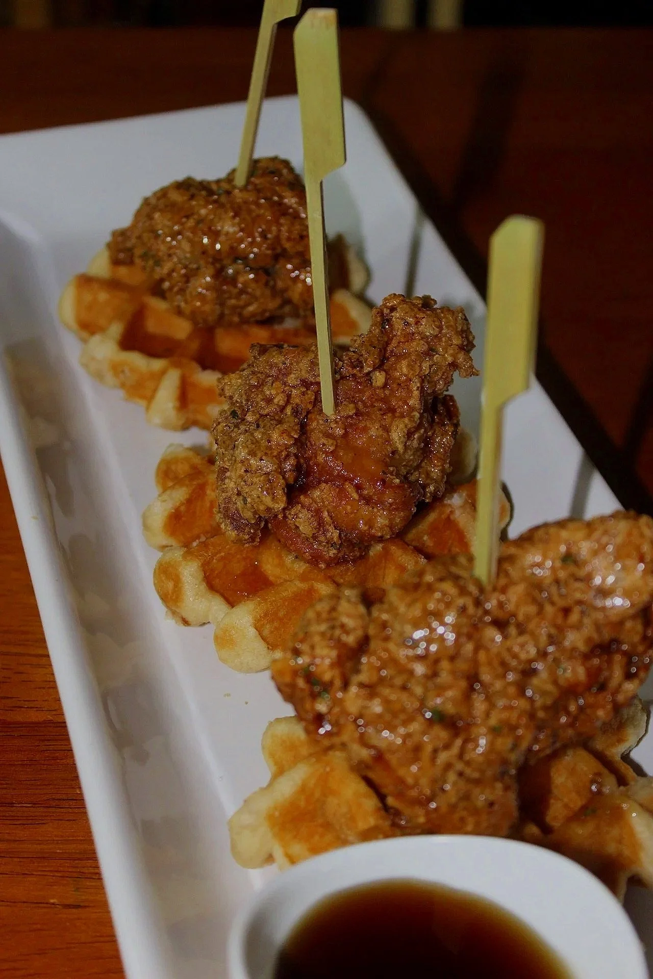 Close-up of fried chicken on a waffle, topped with sauce, served on a white rectangular plate with a small cup of soy sauce on the side.
