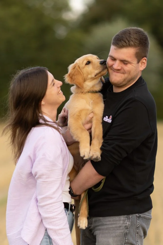 Ein Mann und eine Frau halten gemeinsam einen niedlichen Golden Retriever Welpen. Die Frau trägt einen hellen Pullover, der Mann ein schwarzes Sweatshirt. Sie sind im Freien, umgeben von Bäumen, und lachen glücklich.