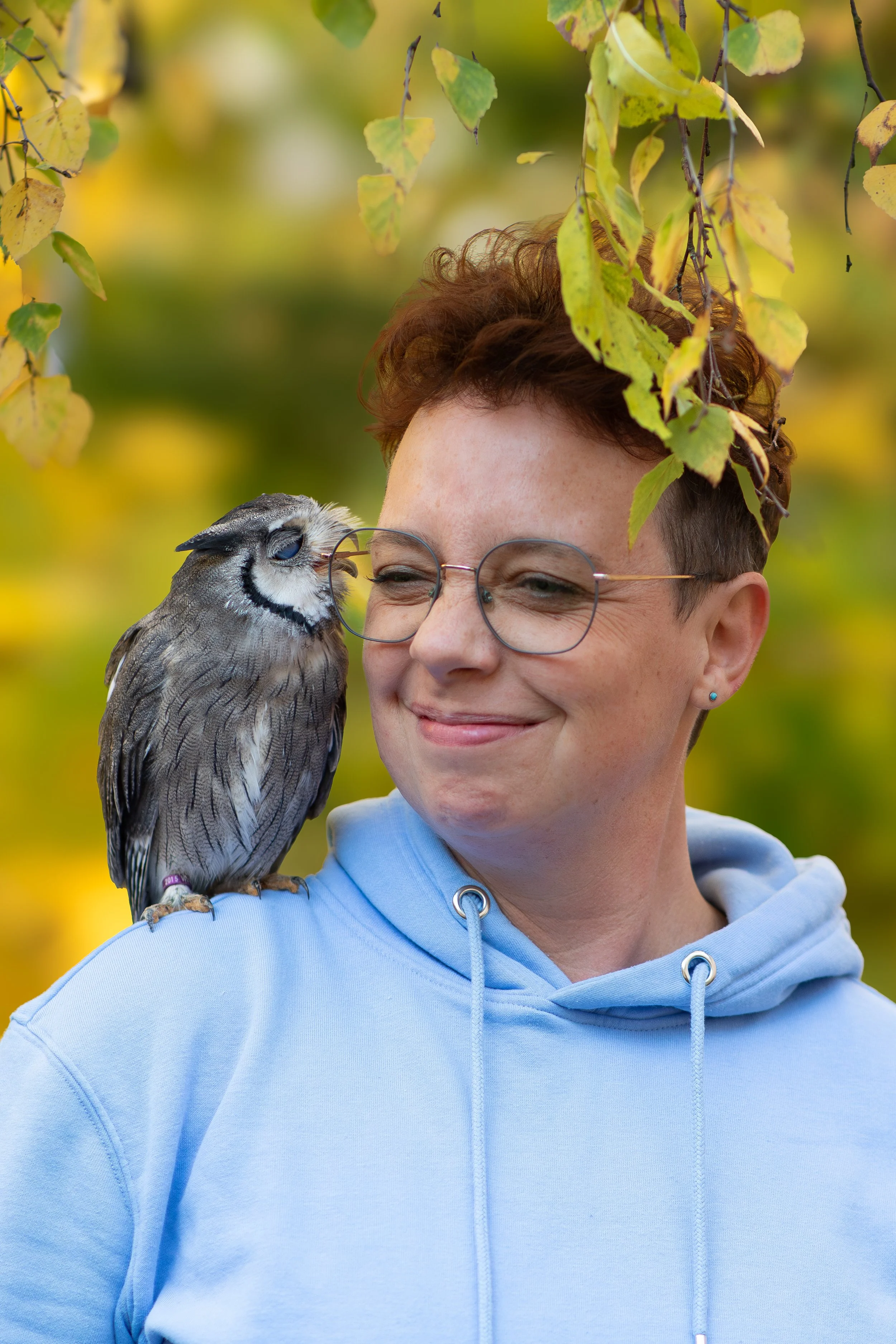 Junge Frau mit kurzen braunen Haaren und runden Brillen, trägt einen hellblauen Hoodie, steht draußen unter einem Baum mit gelben Blättern, hat ein Eulentag am Fuß.