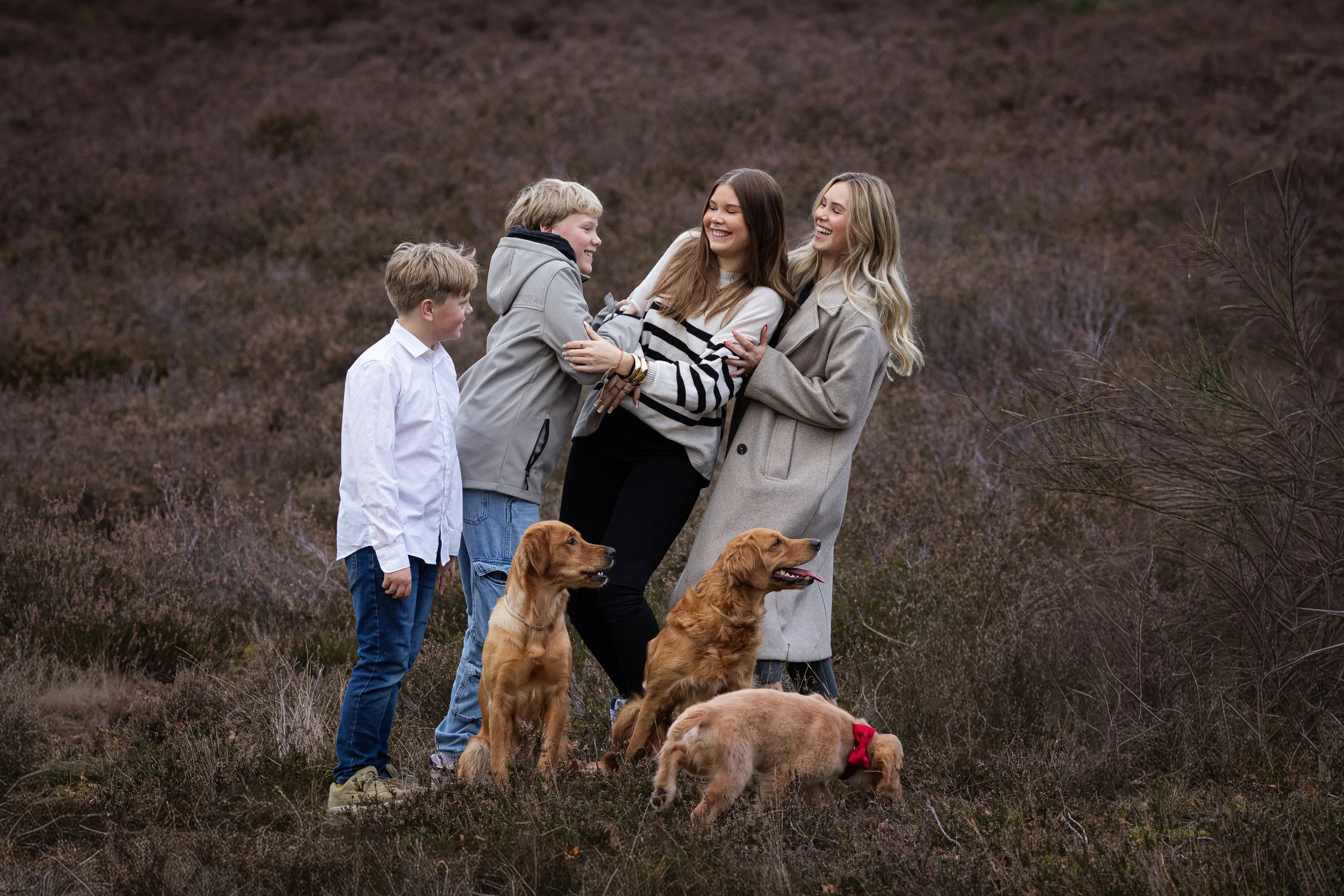 Fünf Menschen, drei Frauen und zwei Jungen, stehen in einer Blumenwiese, umgeben von mehreren Golden Retrievern. Alle lachen und scheinen gemeinsam eine schöne Zeit zu haben.